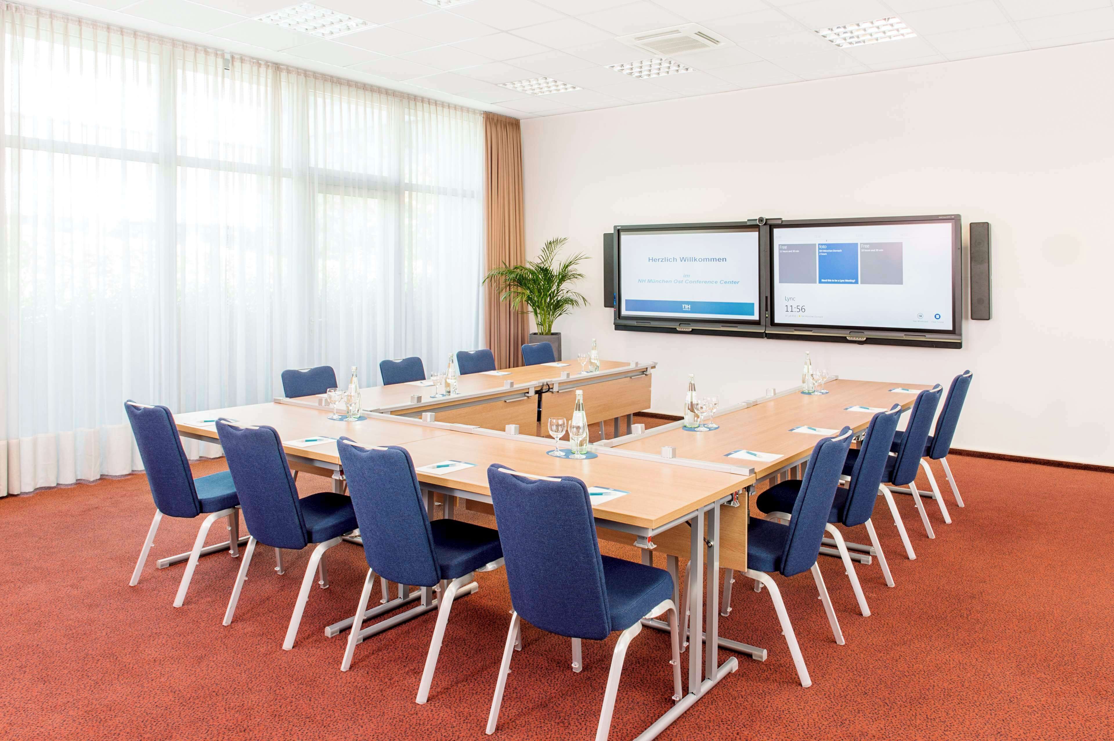 A conference room with a long table, blue chairs, a plant, and two screens displaying information.