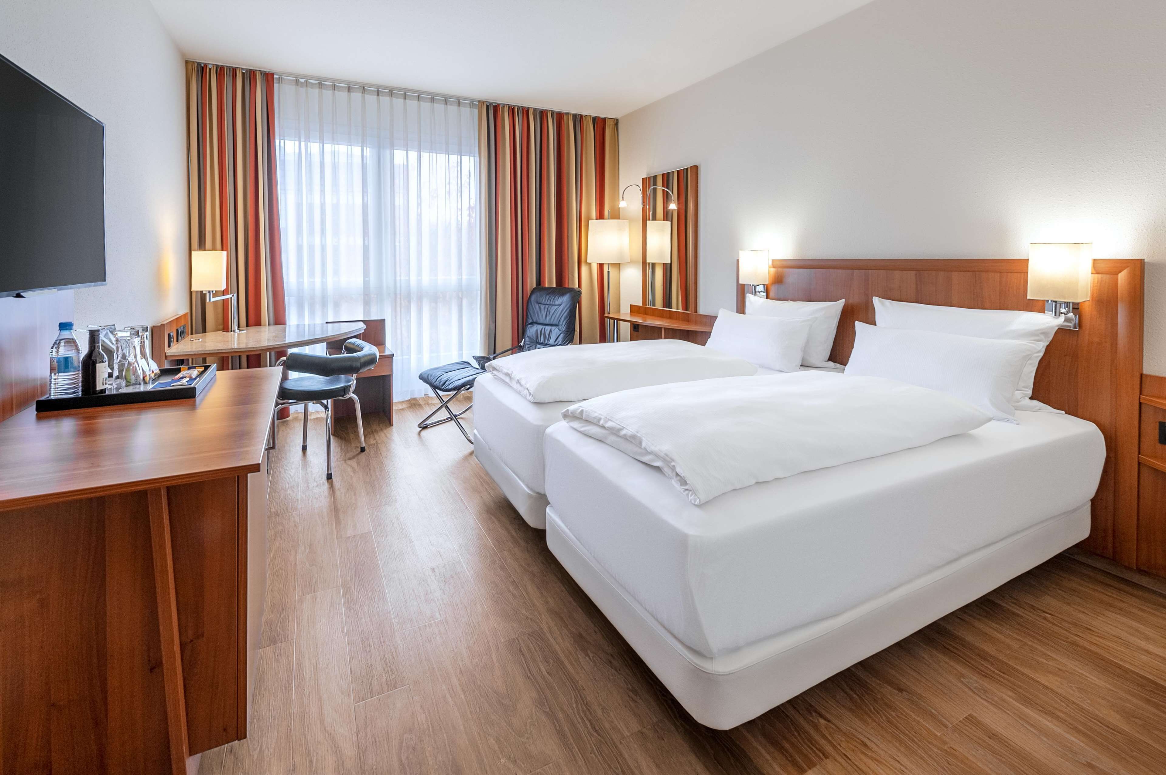 A hotel room with a large bed, a desk with a chair, a TV, and a window with curtains.