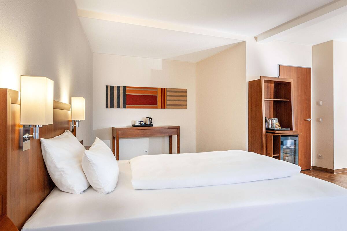 A hotel room with a bed, bedside lamps, a small table with a kettle, and a wardrobe.