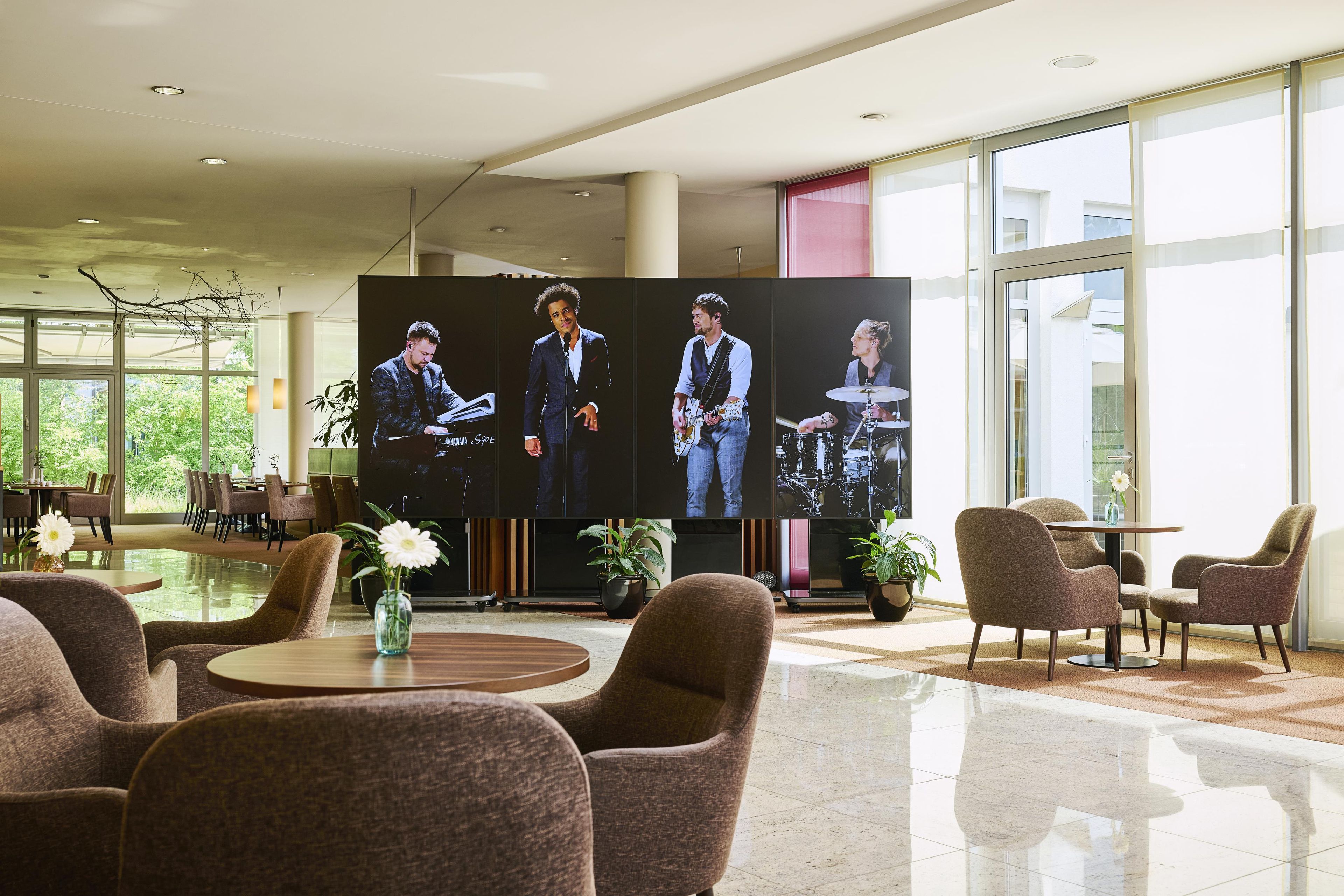 Eine moderne Lobby mit einem großen digitalen Bildschirm, der Bandmitglieder zeigt, bequemen Sitzgelegenheiten und Blick ins Freie.
