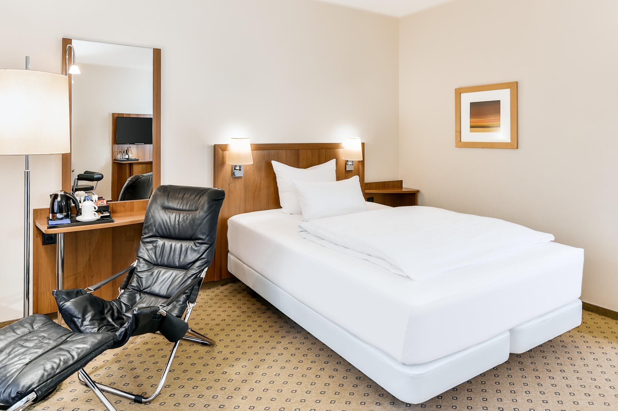 A hotel room with a bed, a chair, a desk with a coffee maker, a mirror, and a picture on the wall.