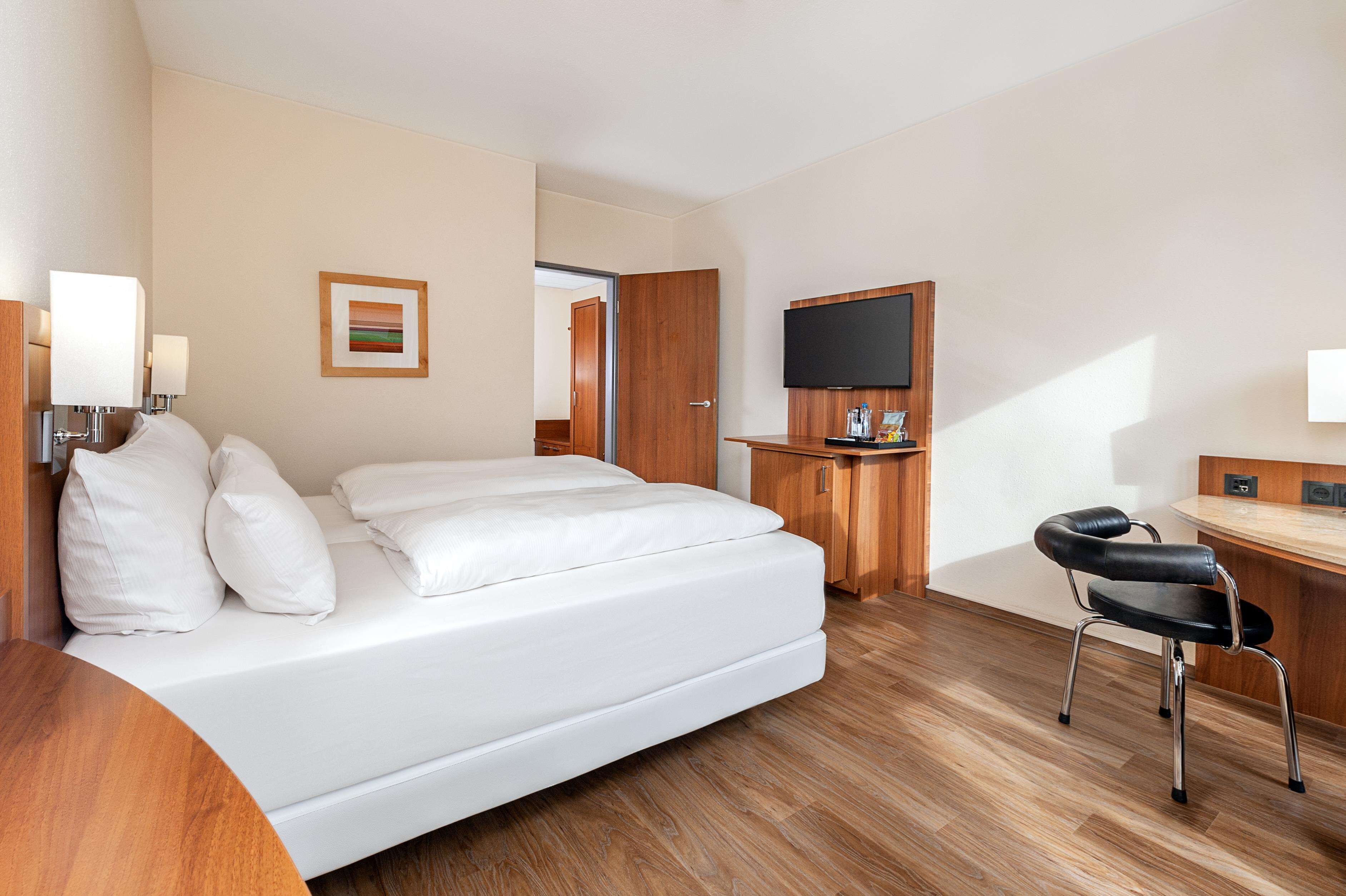 A hotel room with a bed, a desk with a chair, a flat-screen TV, and a wooden door.