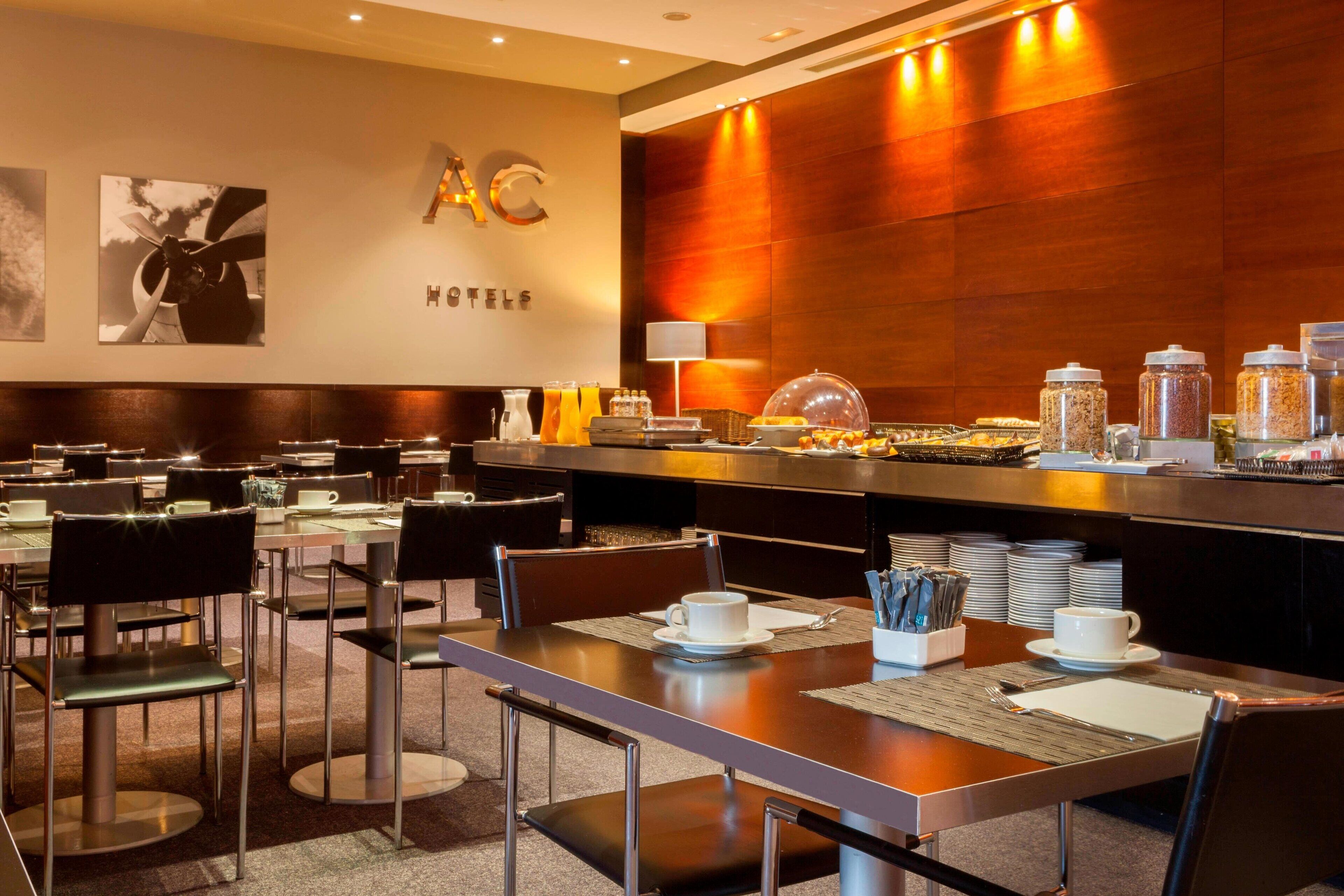 Un área de desayuno de hotel con mesas, sillas y un bufé con recipientes de comida y bebida.