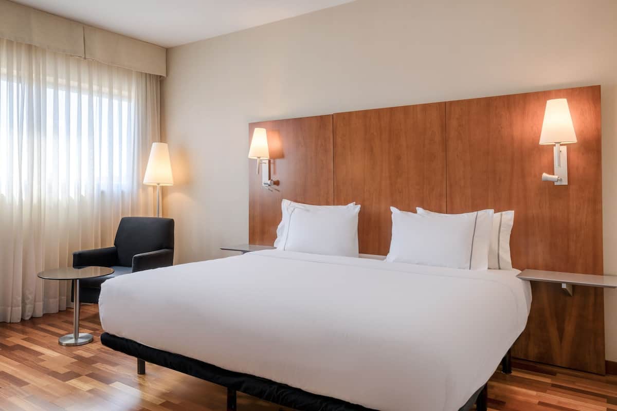 Habitación de hotel con una cama grande, dos lámparas de noche, cabecera de madera, una mesita redonda, una silla y una ventana con cortinas.