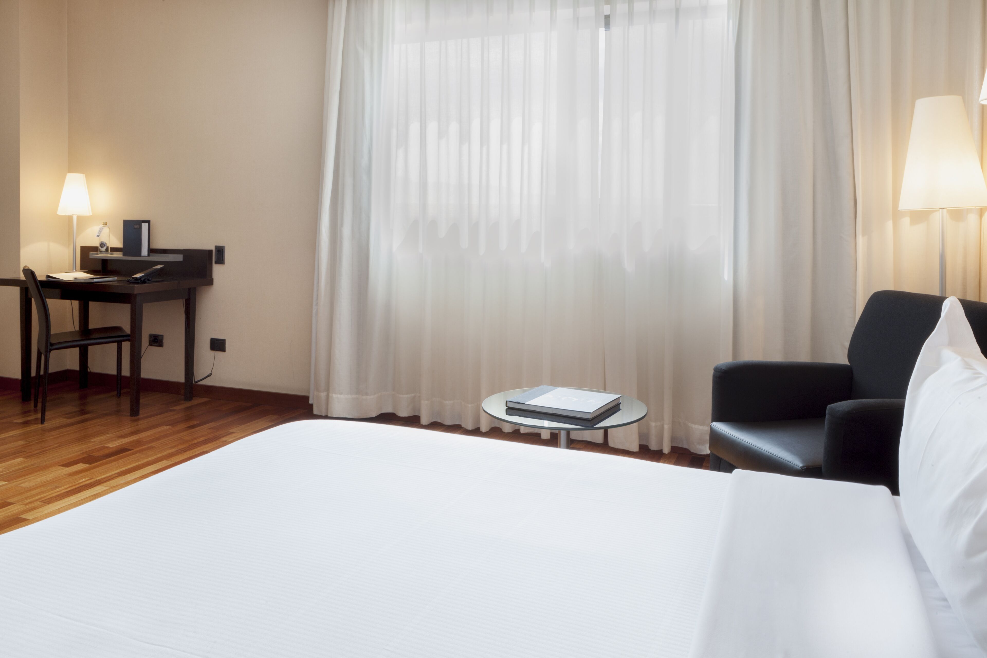 A hotel room with a large bed, a desk with a lamp, a chair, and a small table with a book.