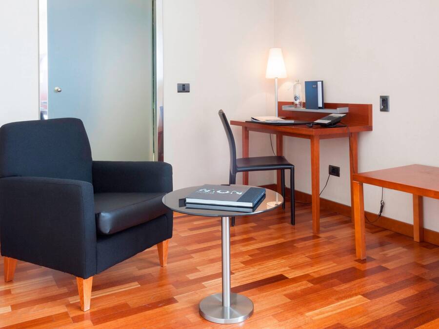 A modern room with a black armchair, a small round table with a book, a wooden desk with a lamp, and a wooden chair.