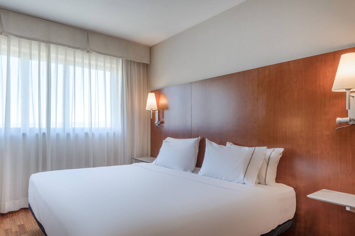 Una cama grande con sábanas blancas, cabecera de madera y dos lámparas de noche.