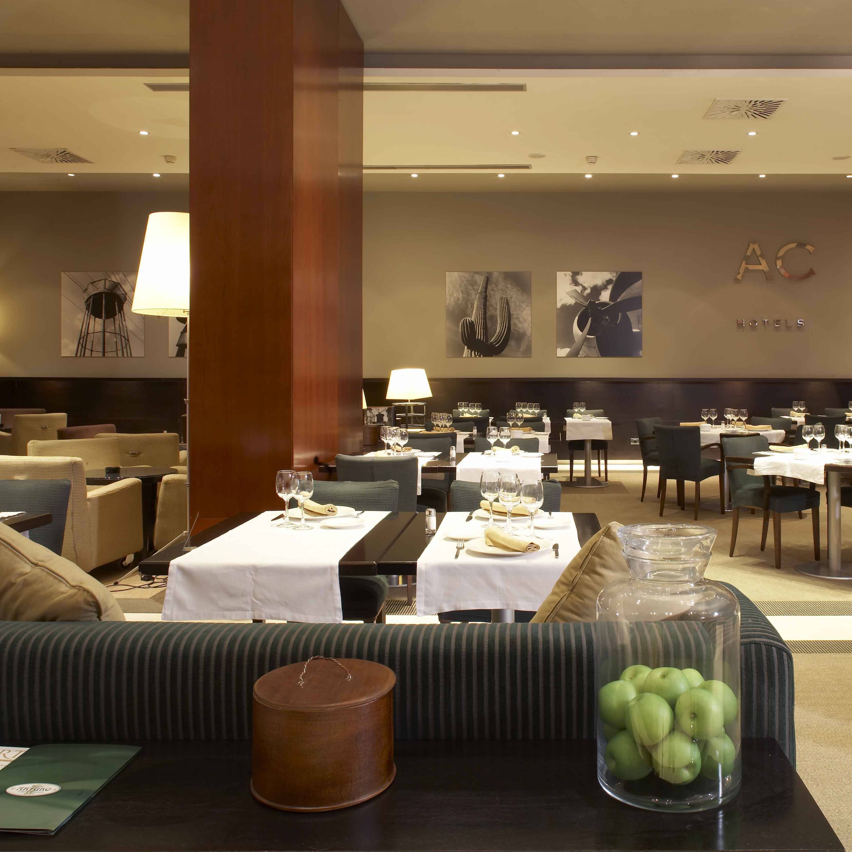 A hotel dining area with tables set for dining, a sofa with a lamp, and a wall with artwork and the hotel name.