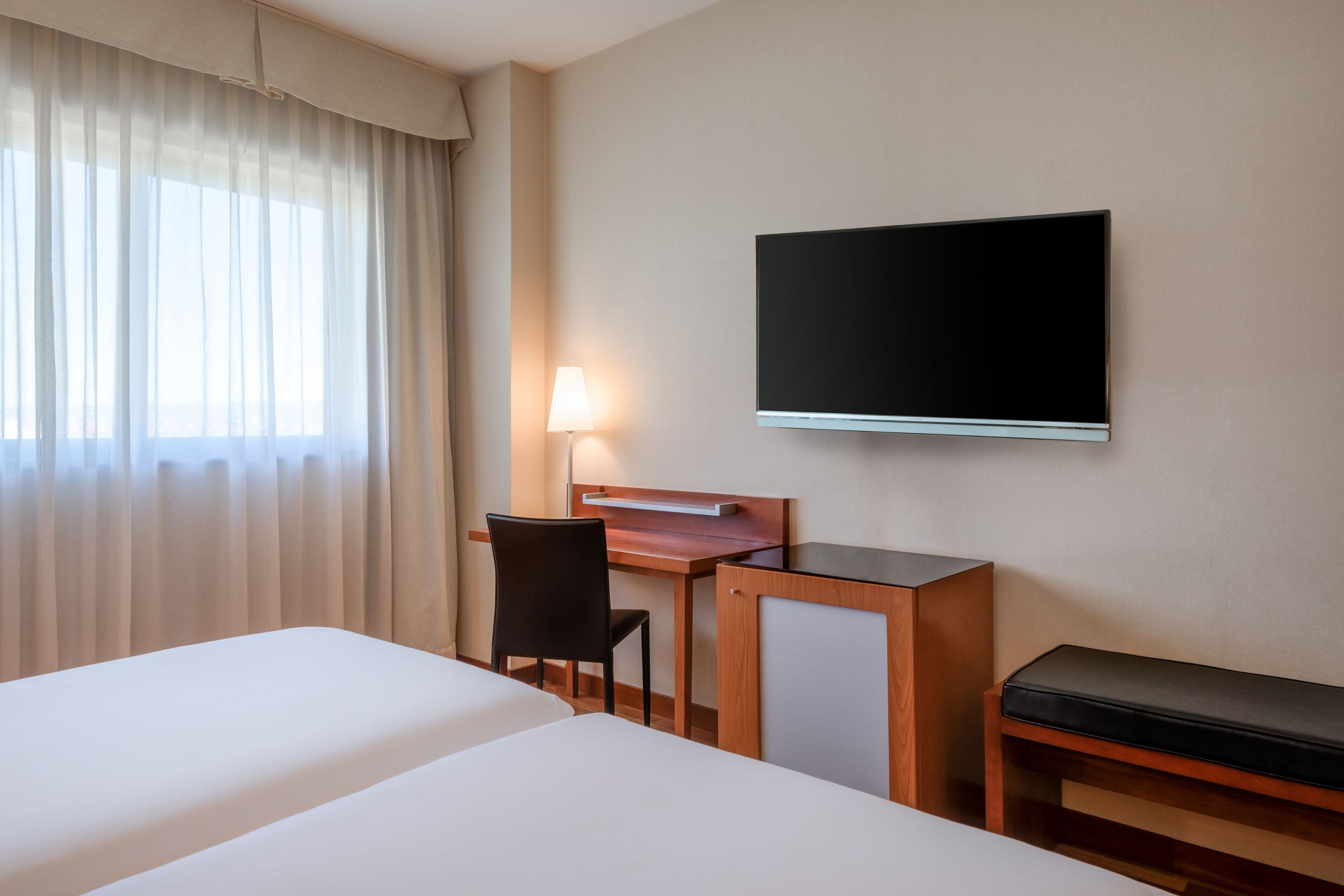 A hotel room with two beds, a desk with a chair, a TV mounted on the wall, and a lamp.