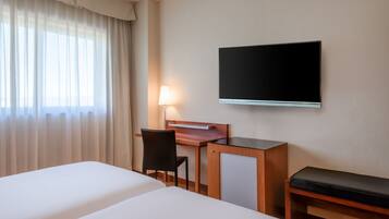 A hotel room with two beds, a desk with a chair, a TV mounted on the wall, and a lamp.