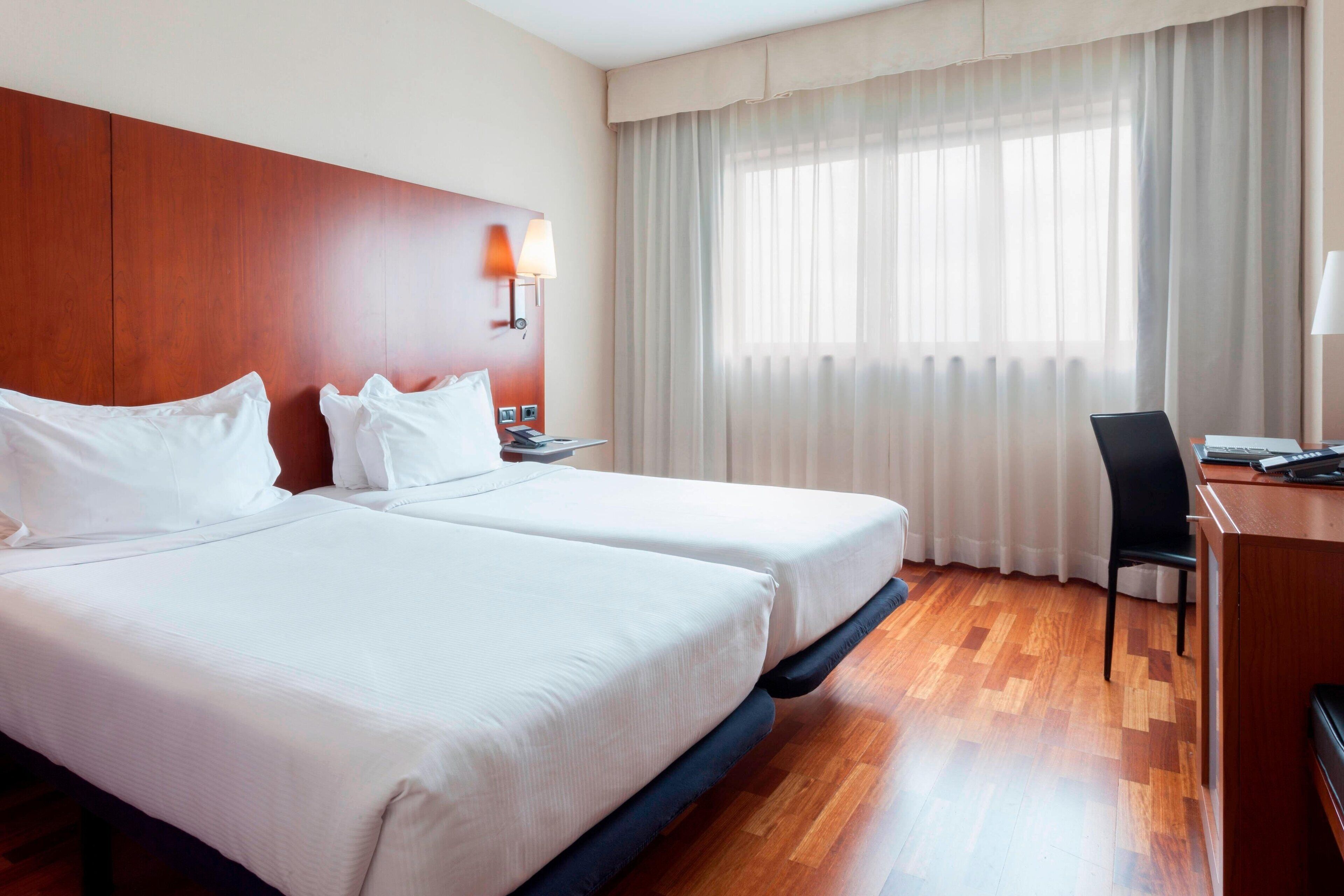 Une chambre d’hôtel avec deux lits, une tête de lit en bois, un bureau avec une chaise et une grande fenêtre avec des rideaux légers.