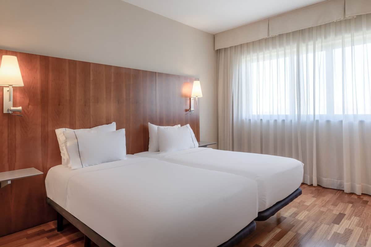 Habitación de hotel con dos camas, cabecero de madera, lámparas de noche, mesitas de noche y un ventanal con cortinas de gasa.