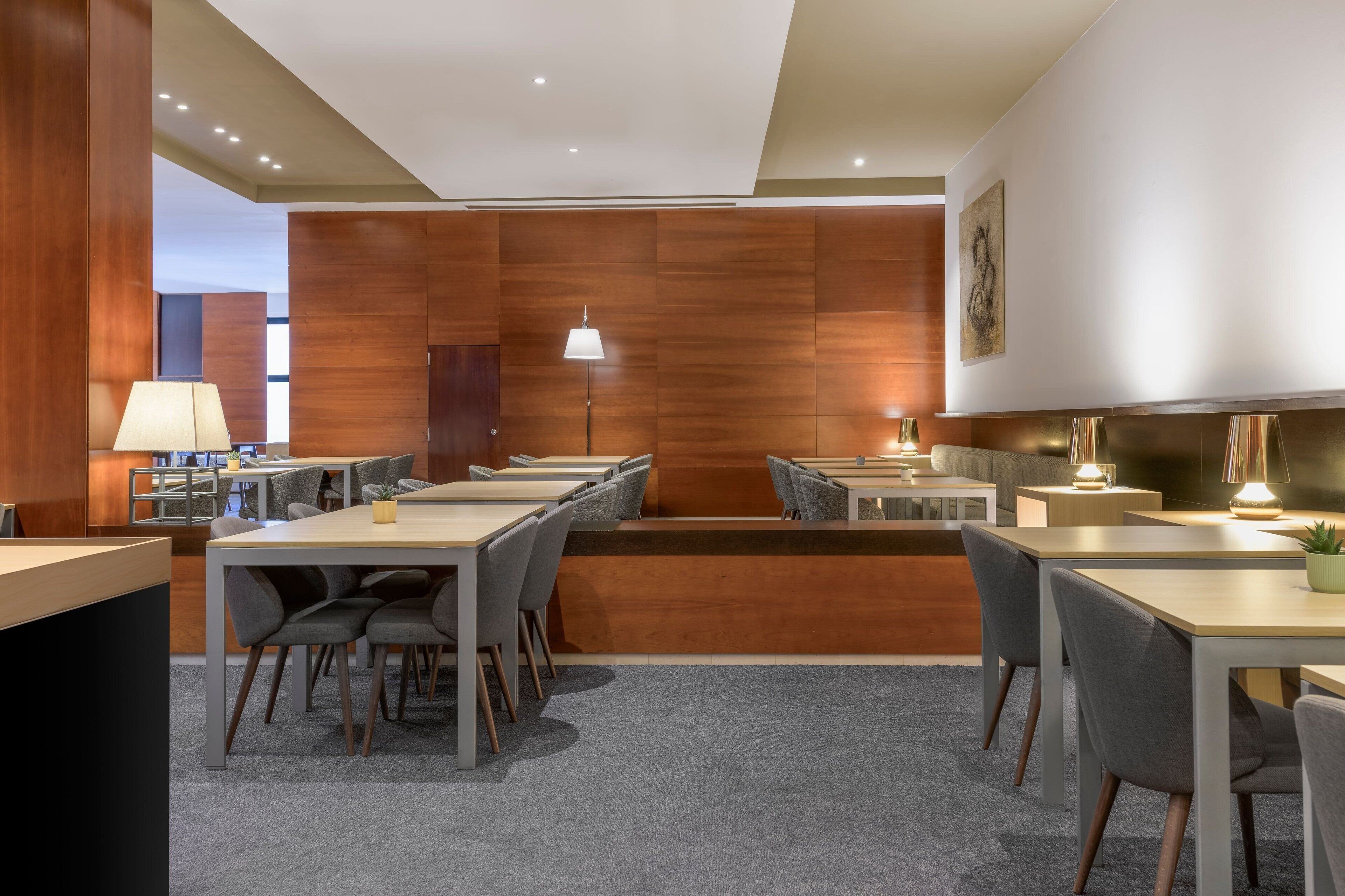Un moderno vestíbulo de hotel con paneles de madera, varias mesas de comedor con sillas y una escalera central.