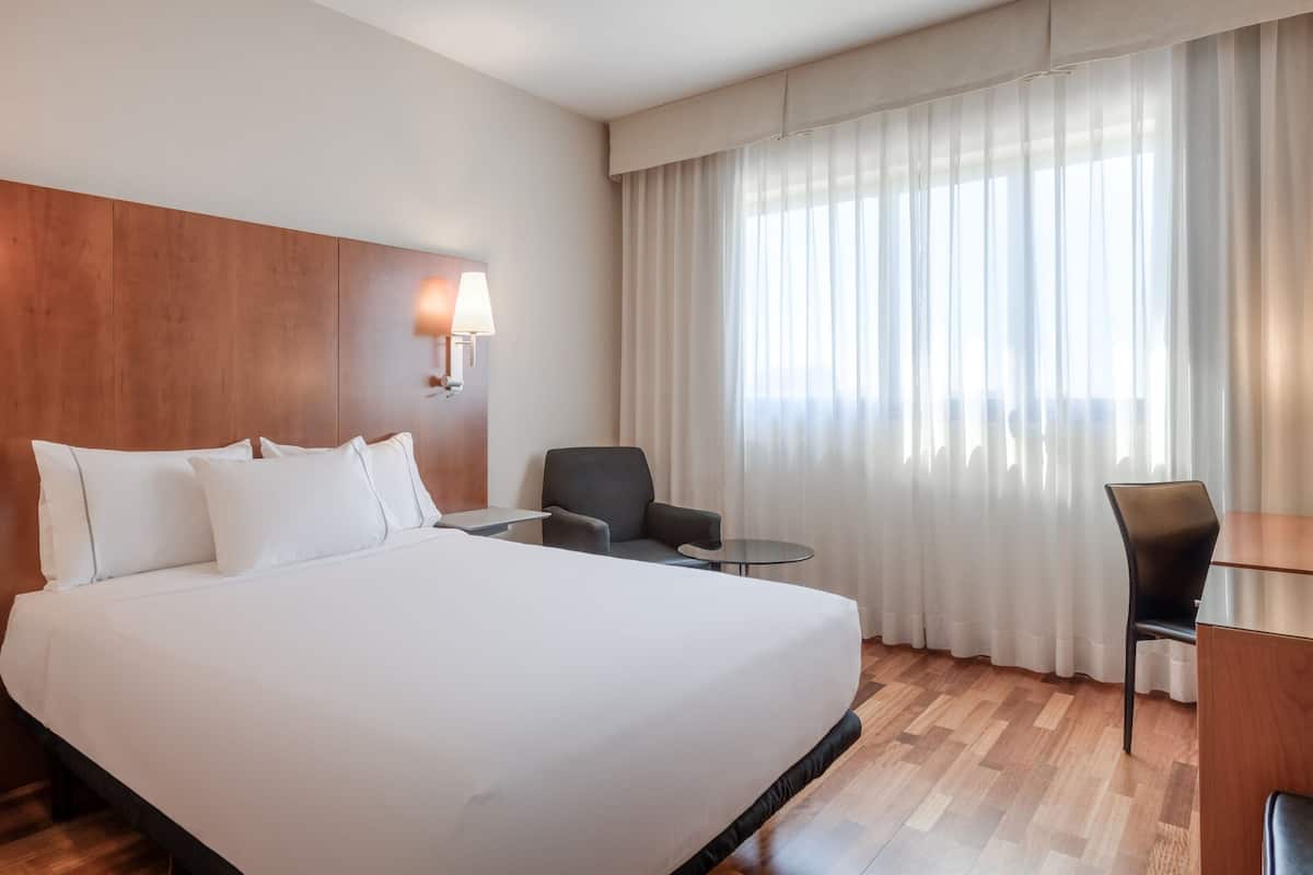 Habitación de hotel con una cama grande, un escritorio, dos sillas, una lámpara y una ventana con cortinas.