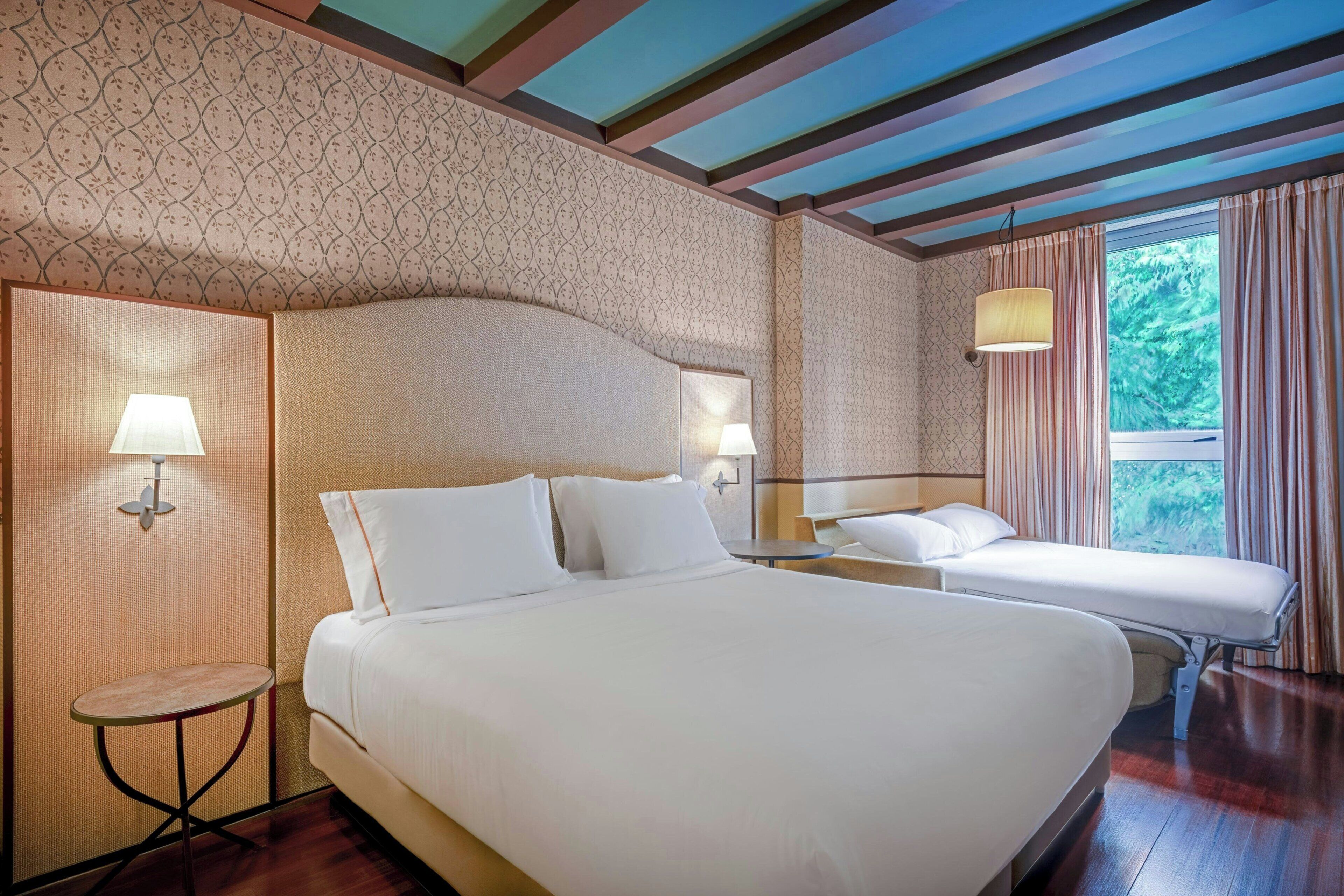 Une chambre d’hôtel avec un grand lit, des tables de chevet, des lampes et une vue sur la verdure par la fenêtre.