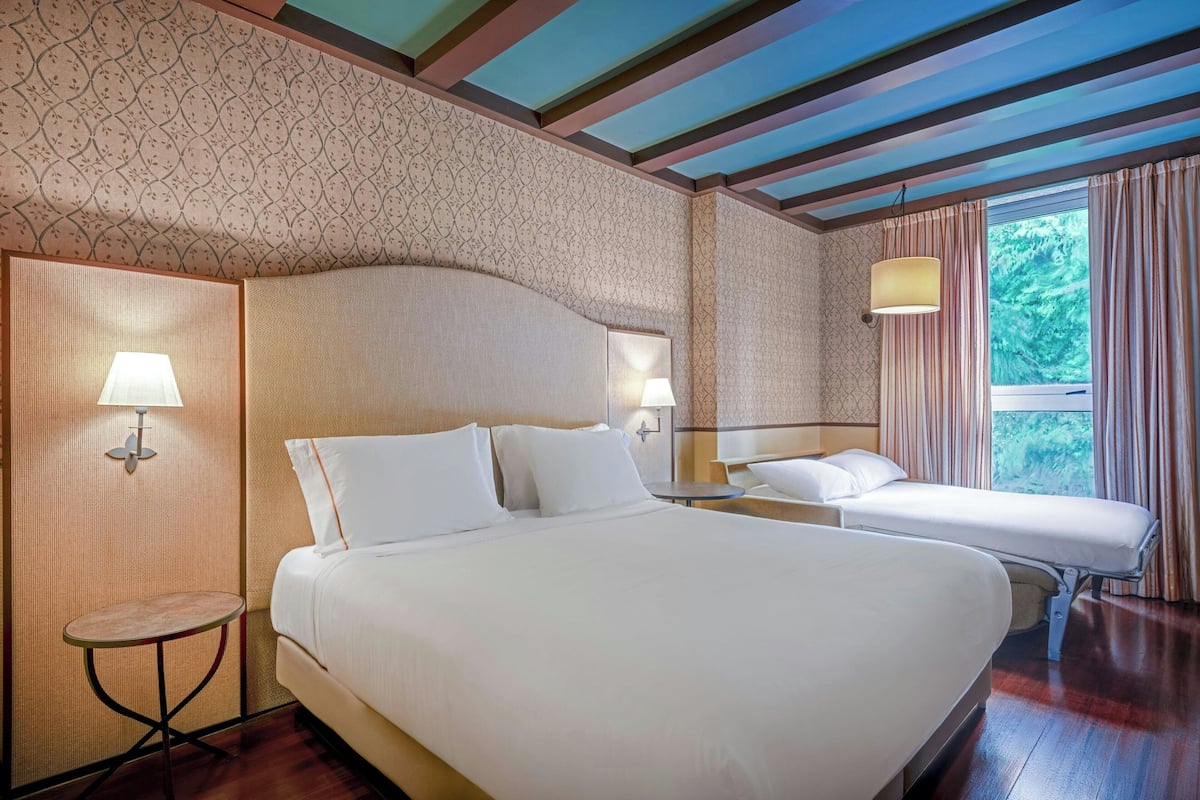 Quarto de hotel com cama grande, mesas de cabeceira, luminárias e vista para o verde pela janela.