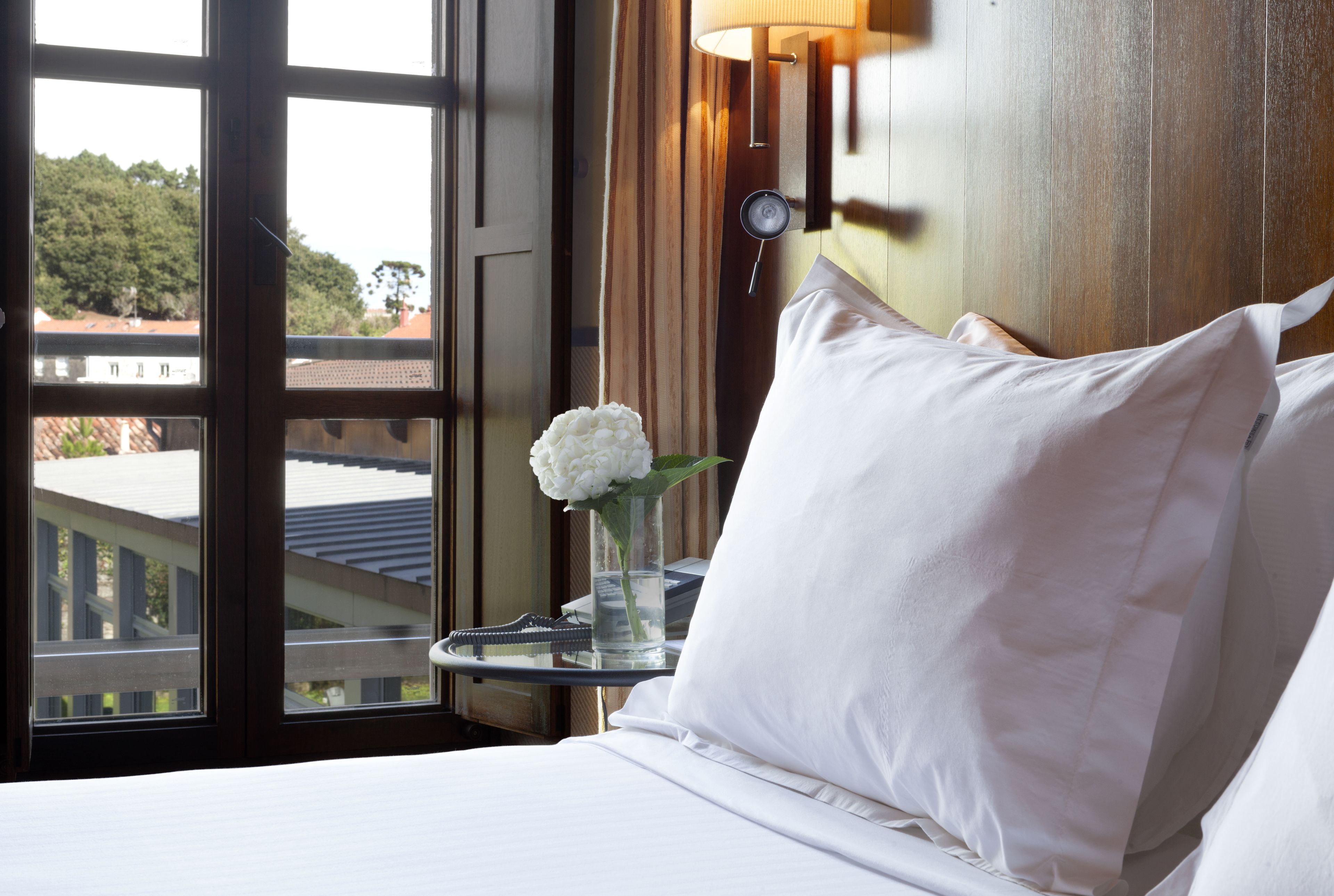 A hotel room with a bed, a bedside table with a vase of flowers, a lamp, and a view of buildings through a window.