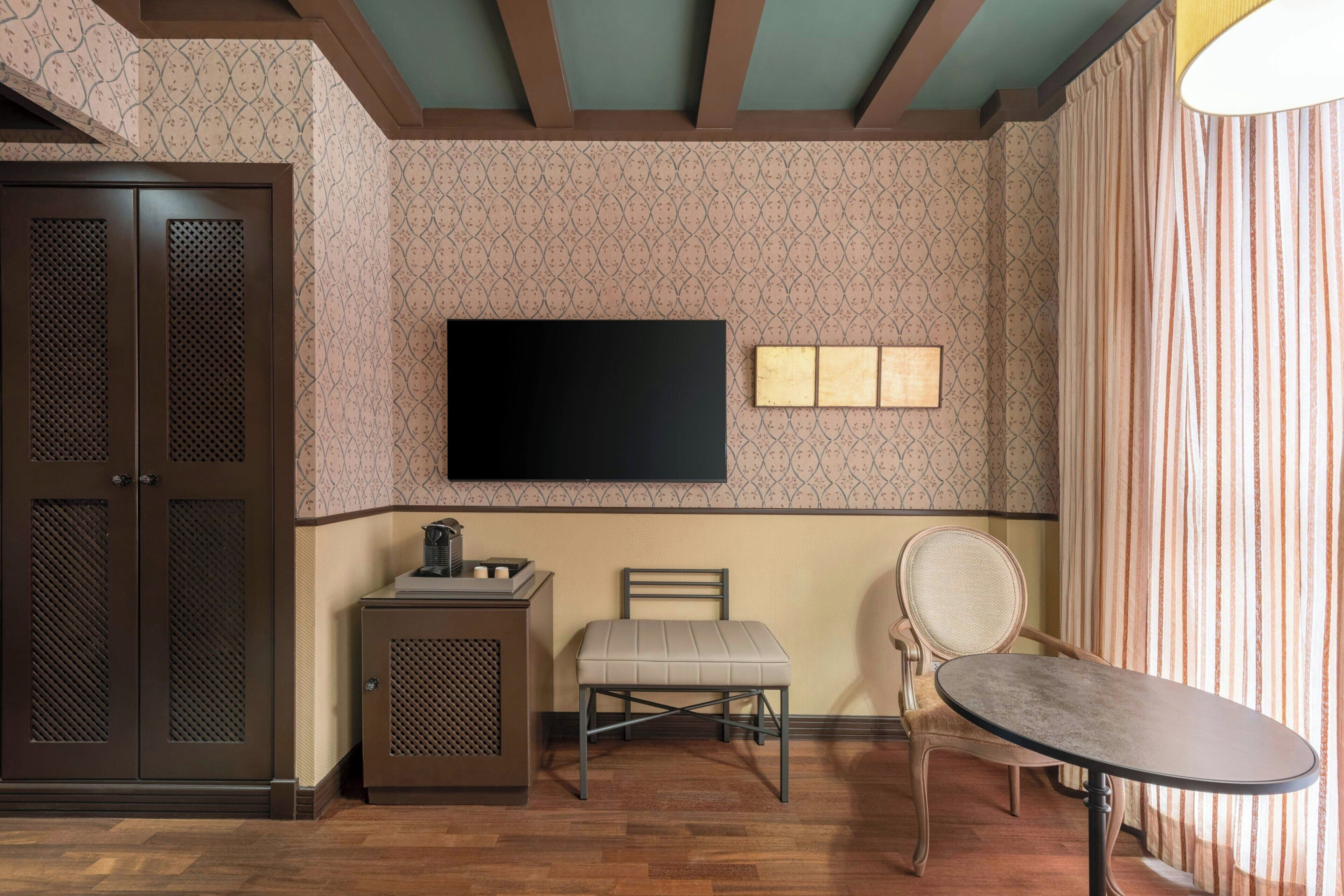 Quarto com piso de madeira, televisão na parede, um pequeno armário, um banquinho acolchoado, uma cadeira e uma mesa.