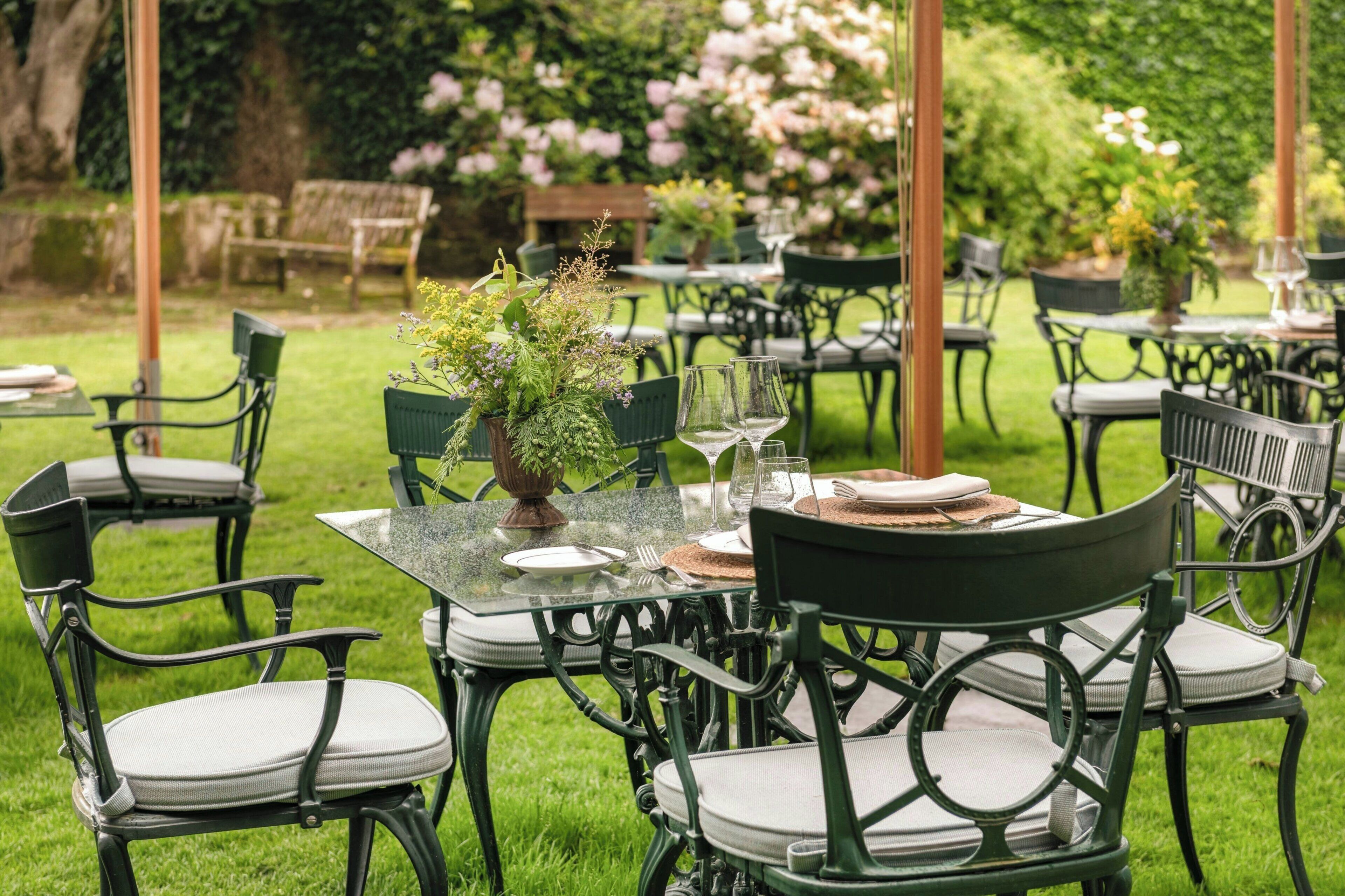 An outdoor dining area with green metal chairs and tables, surrounded by lush greenery and flowers.