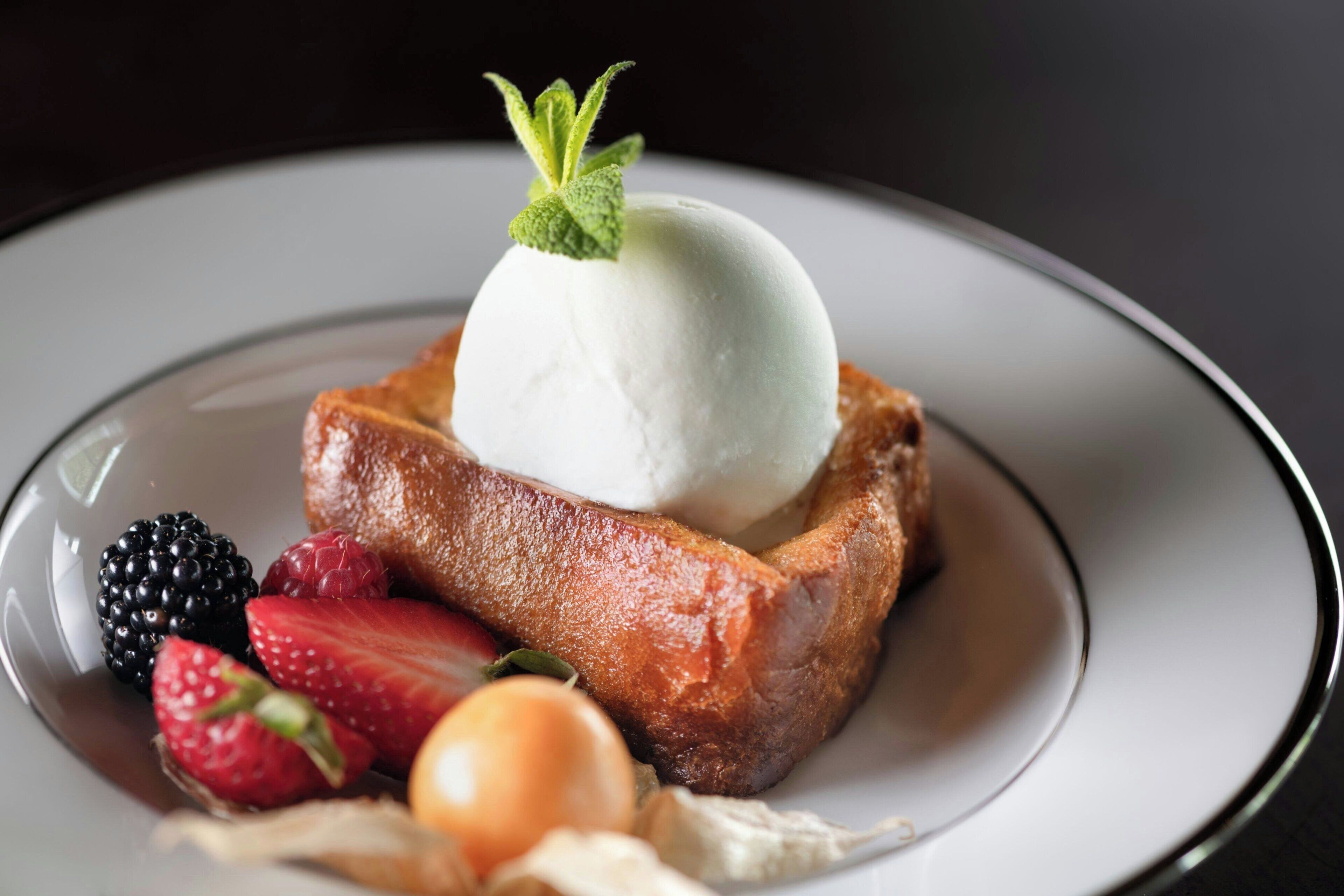 A dessert plate with a slice of toast topped with a scoop of ice cream and garnished with a mint leaf, accompanied by strawberries, a blackberry, and a cherry.