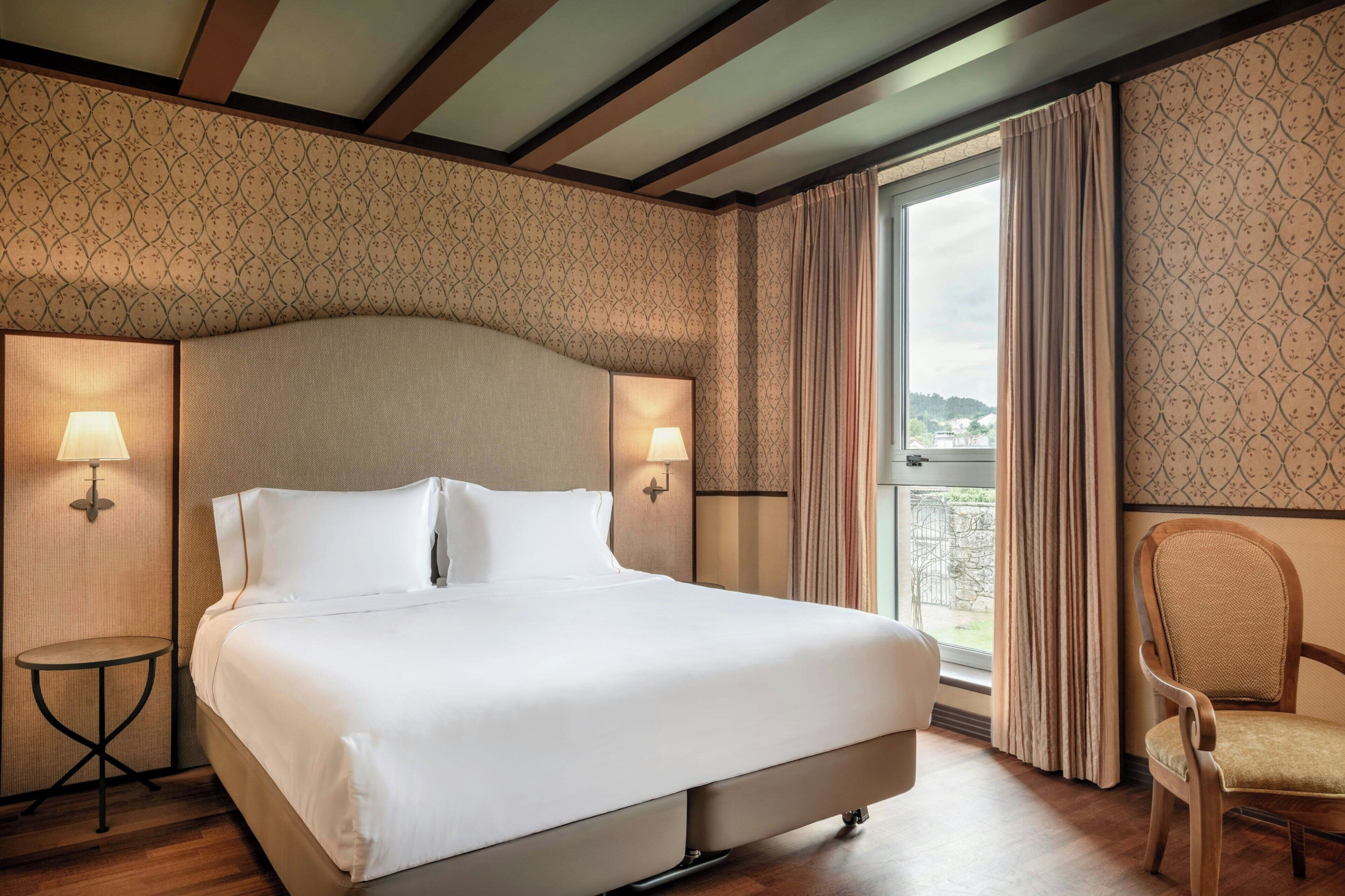 A hotel room with a large bed, two wall-mounted lamps, a small round table, a chair, and a view through a window with curtains.