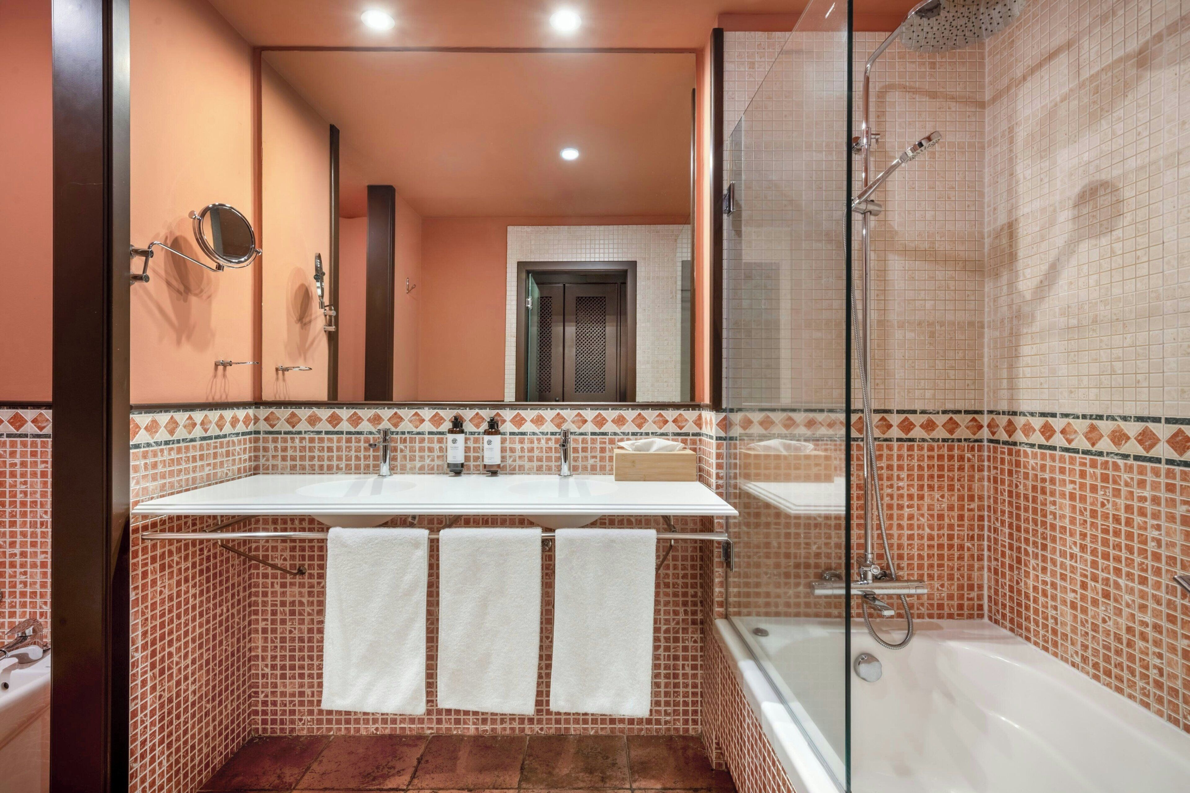 Un baño con cabina de ducha de vidrio, un lavabo blanco con dos espejos y suelo de baldosas.
