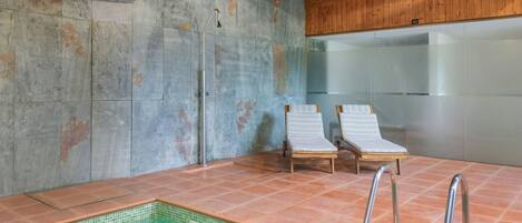 A modern indoor pool area with a glass wall, two lounge chairs, and a metal ladder.