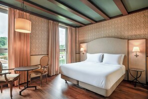 A hotel room with a large bed, two bedside lamps, a small table, a chair, and a view of the outdoors through a window.