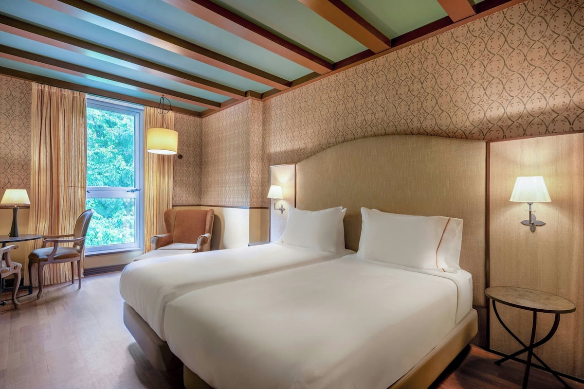 Quarto de hotel com duas camas, uma escrivaninha, uma cadeira e uma janela com cortinas.