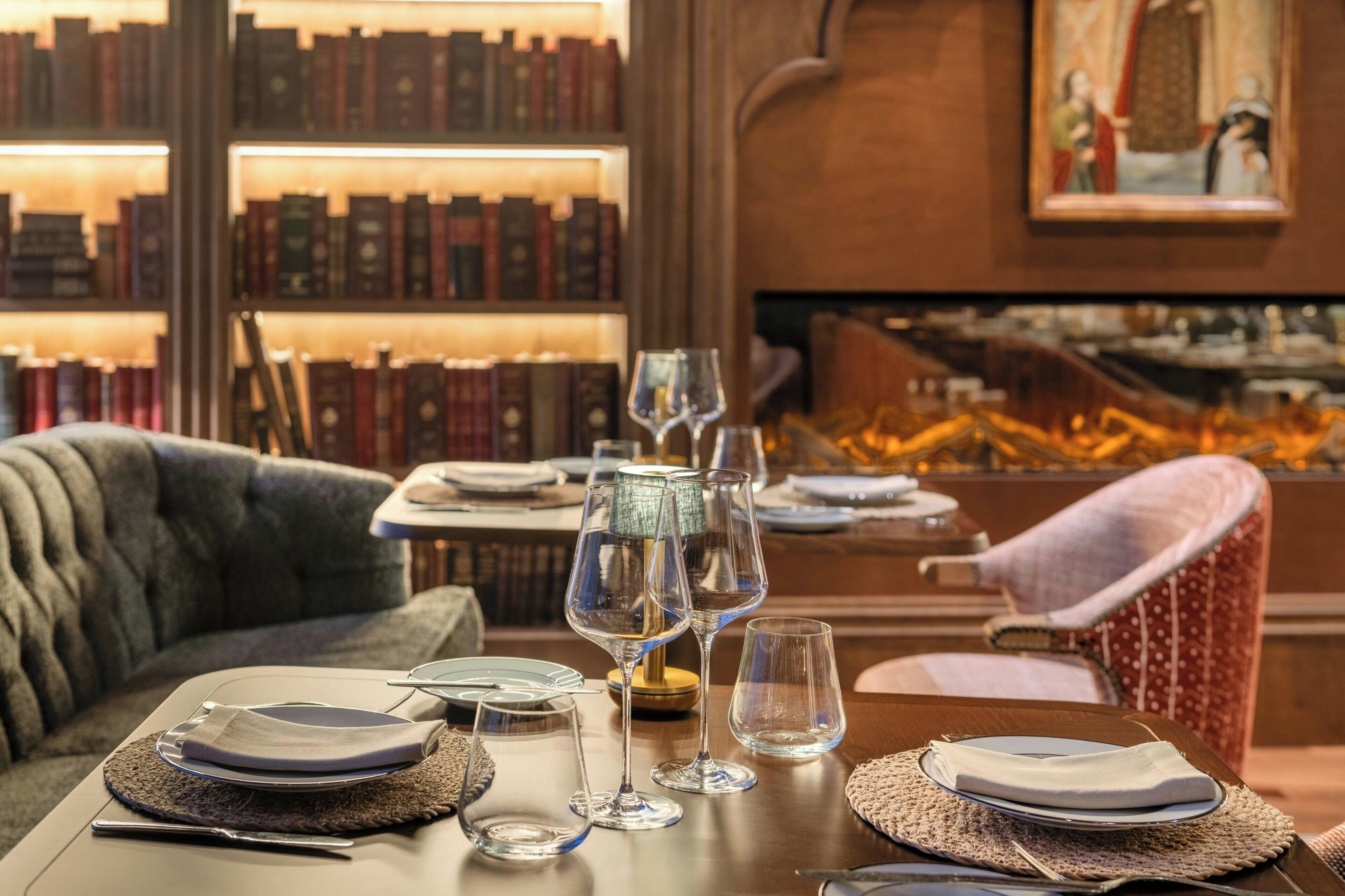 A dining table set with plates, cutlery, and wine glasses, surrounded by bookshelves and a patterned armchair.