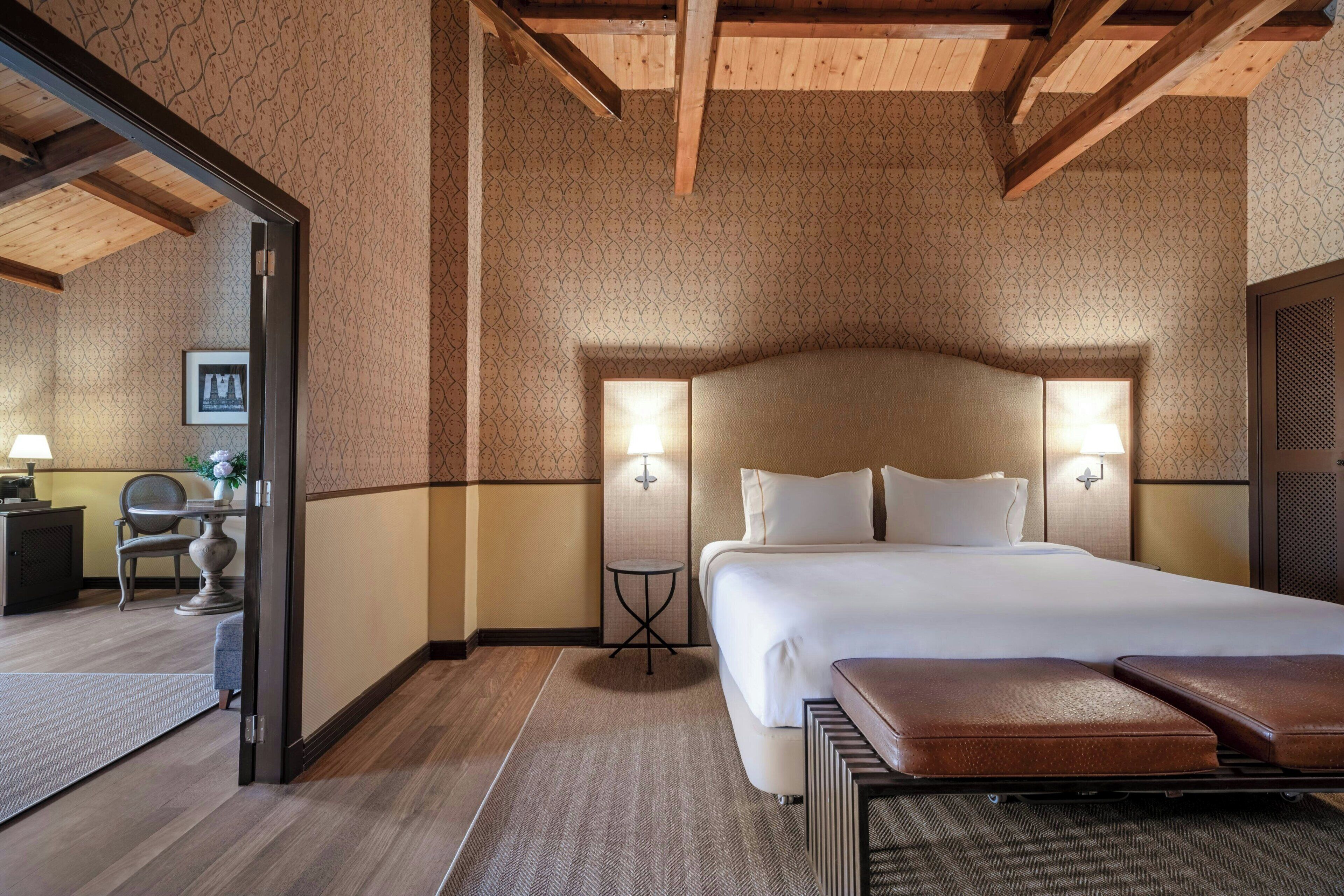 A hotel room with a large bed, two bedside lamps, a wooden ceiling, and a view of the adjacent room.