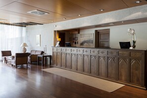 A reception area with a wooden reception desk, a seating area with chairs, a lamp, and a vase with flowers. - Hotel Palacio del Carmen, Autograph Collection (Santiago de Compostela)
