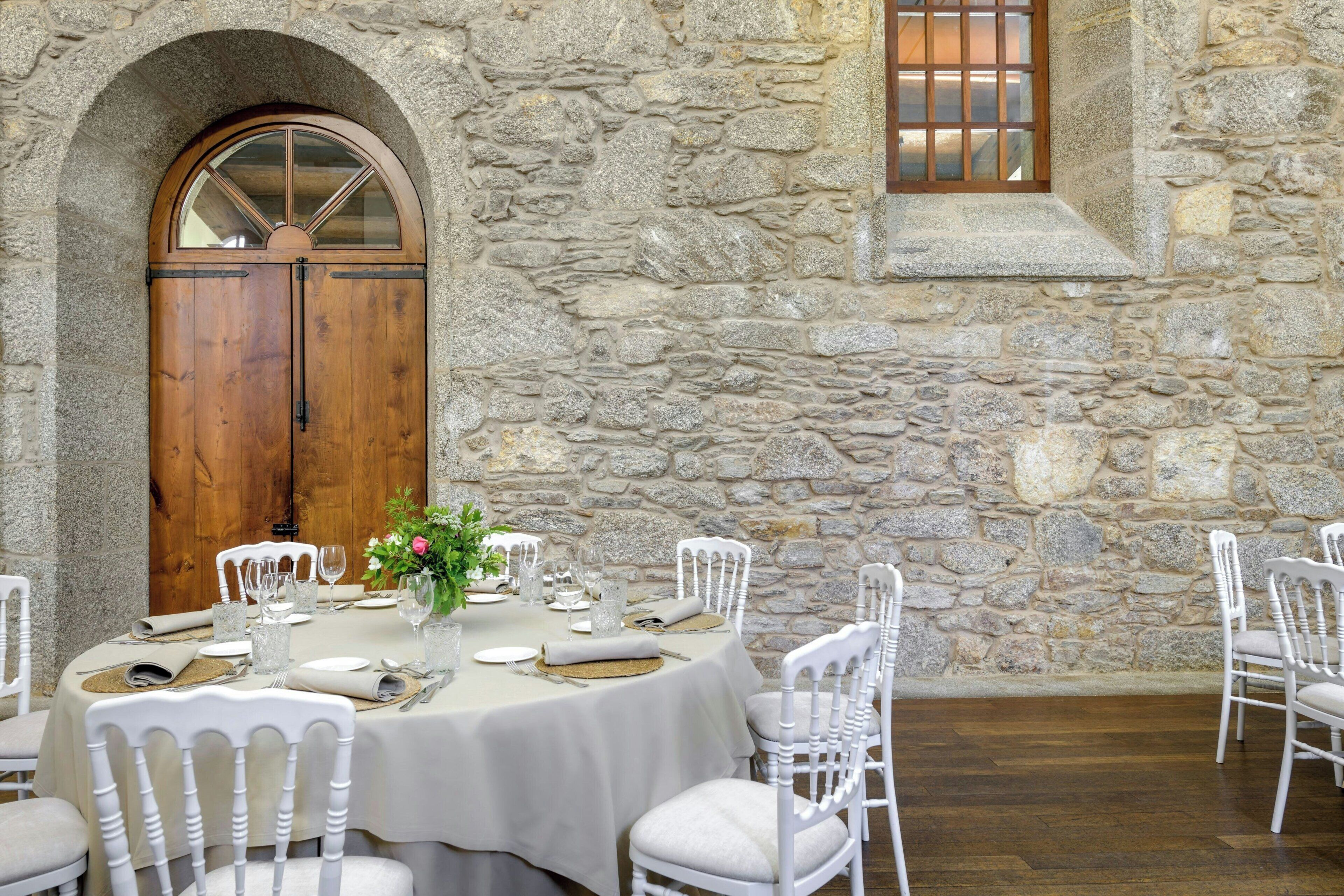 Comedor de paredes de piedra con una mesa redonda para cuatro, sillas blancas y una puerta de madera con panel de vidrio.
