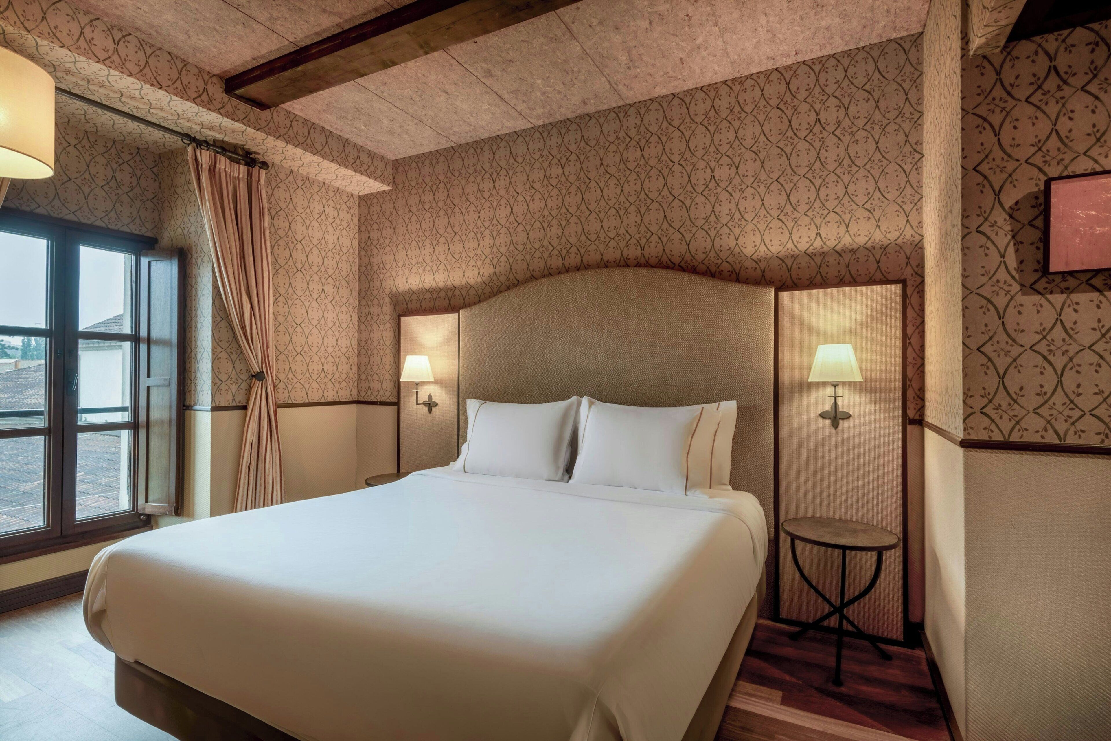 Una habitación de hotel con una cama grande, dos lámparas de noche, una mesita y una ventana con cortinas.