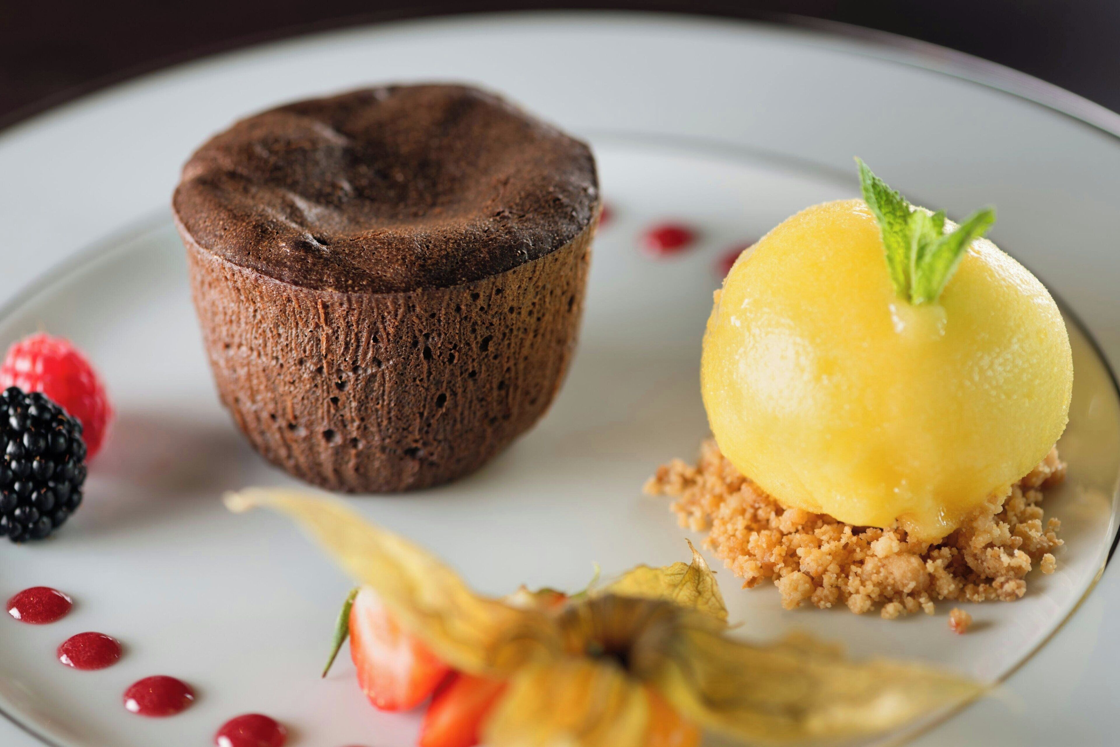 Un plato de postre con pastel de chocolate y cobertura de fruta amarilla, acompañado de bayas y una hoja decorativa.