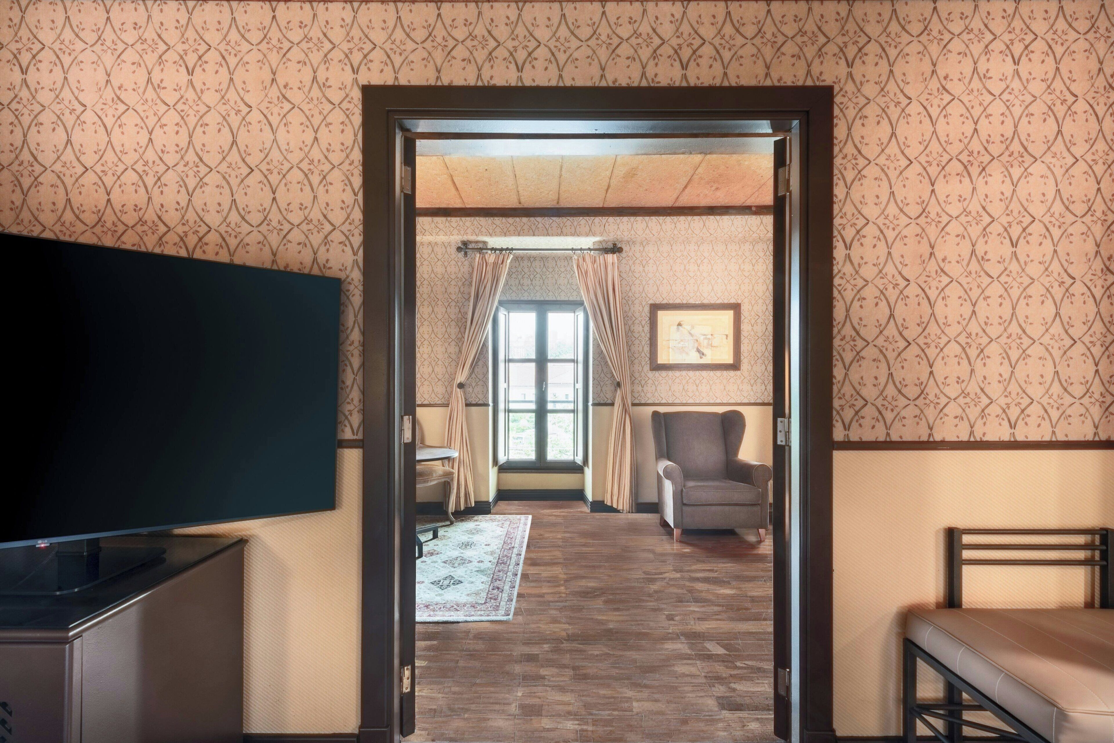 A room with a television, a bed, a chair, a window with curtains, and a patterned wall.