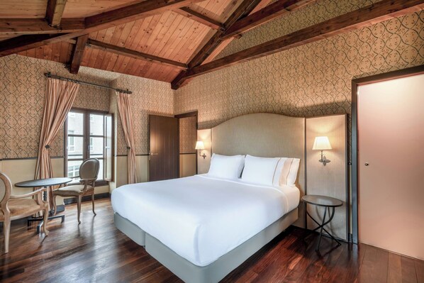 A hotel room with a large bed, two bedside lamps, a small round table, a chair, and a window with curtains.