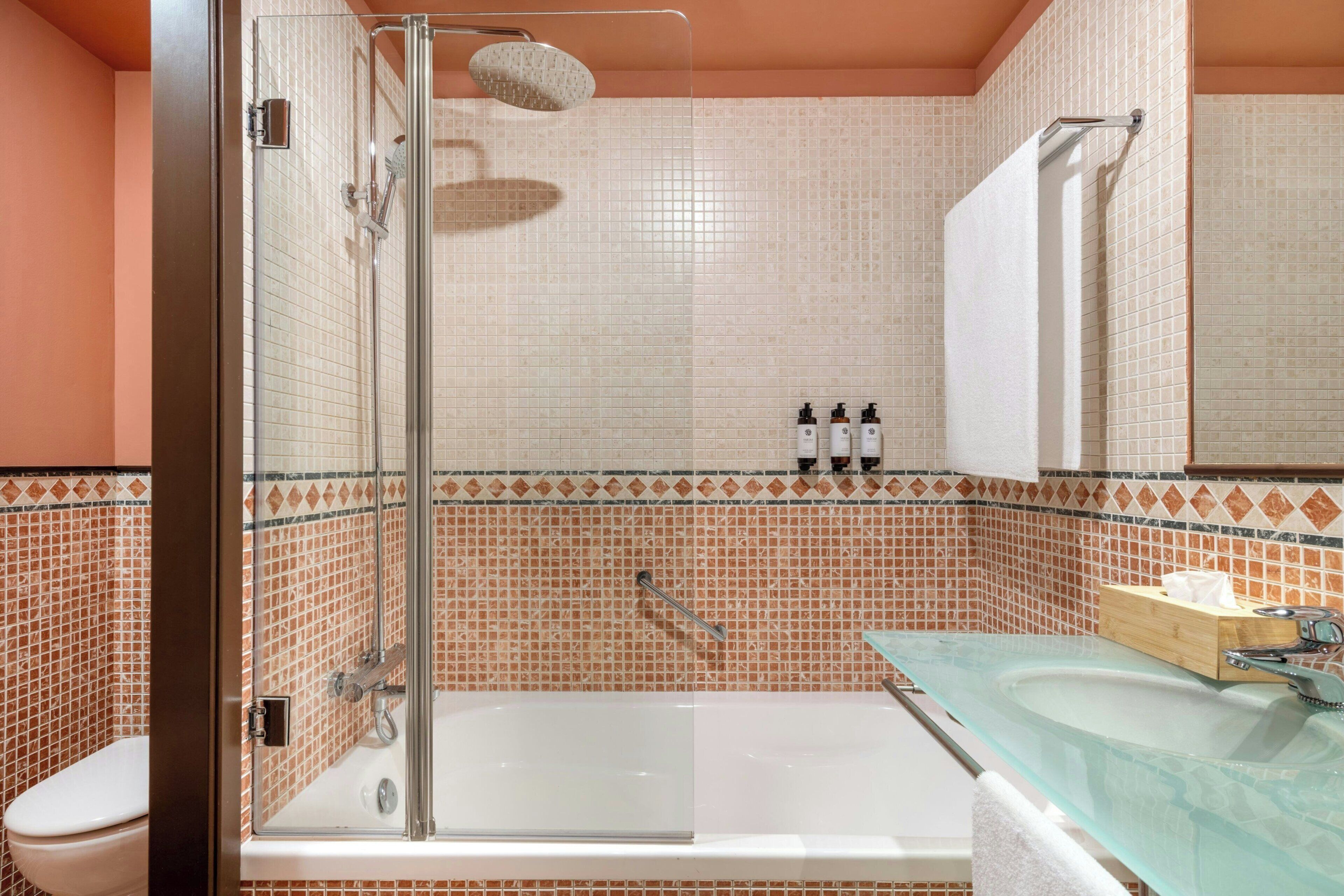 Een badkamer met een douche met glazen wand, een bad, een wastafel met glazen blad en een spiegel boven de wastafel.
