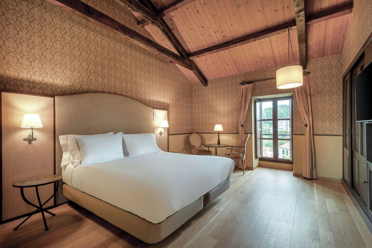Quarto de hotel com uma cama grande, duas luminárias de cabeceira, uma mesinha, uma cadeira e uma janela com cortinas.