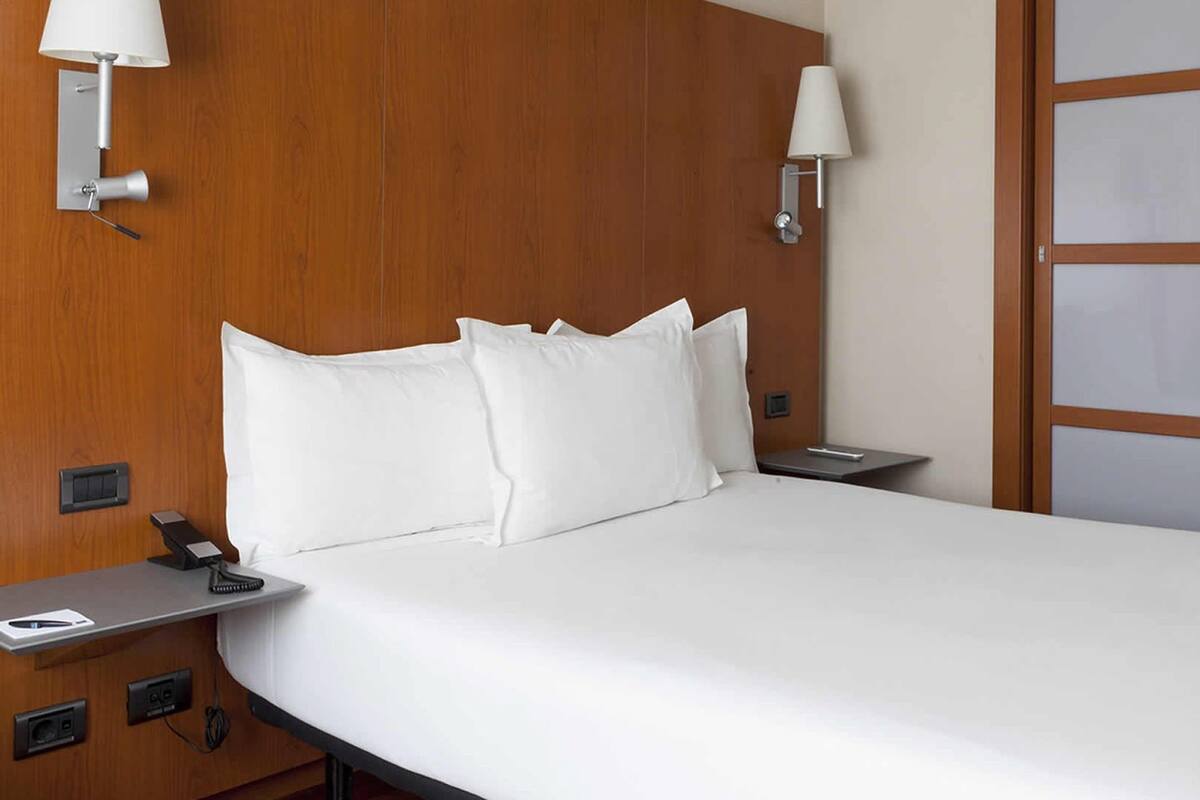 Una habitación de hotel con cabecera de madera, cama con ropa de cama blanca, mesita de noche con lámpara y teléfono.