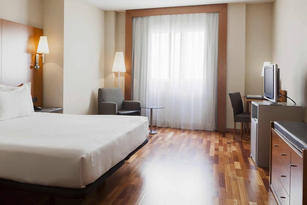 Habitación de hotel con una cama grande, dos lámparas de noche, un escritorio con una silla, un televisor y una ventana con cortinas.
