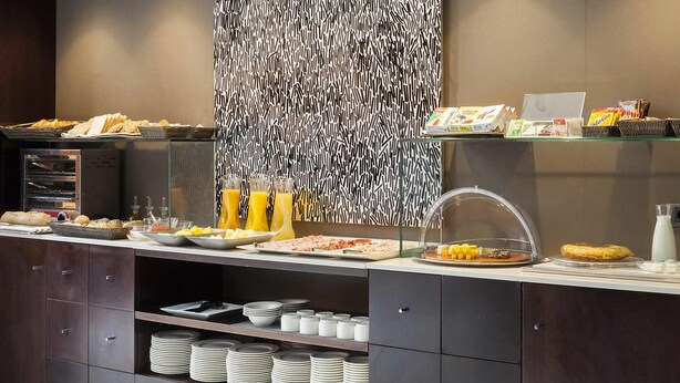 A buffet setup with various food items, including bread, pastries, and beverages, with a modern patterned wall and neatly arranged plates and bowls. - Hotel Ciudad de Lleida (Lleida)