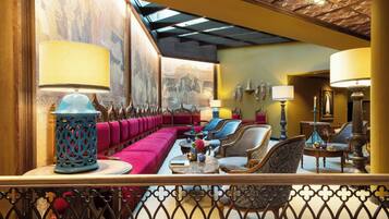 A spacious lounge area with a long red sofa, multiple chairs, and tables. There are decorative lamps and wall art.