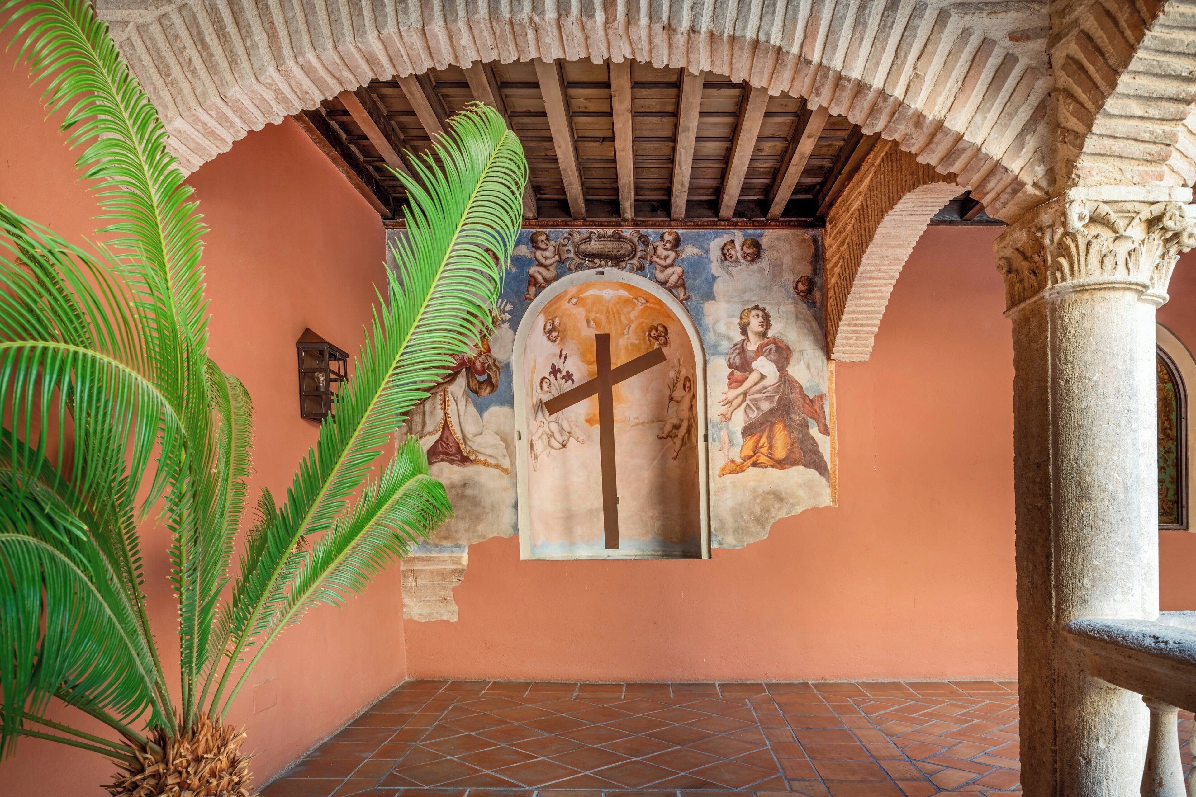 A room with a tiled floor, a large plant, and a mural with a cross and figures.