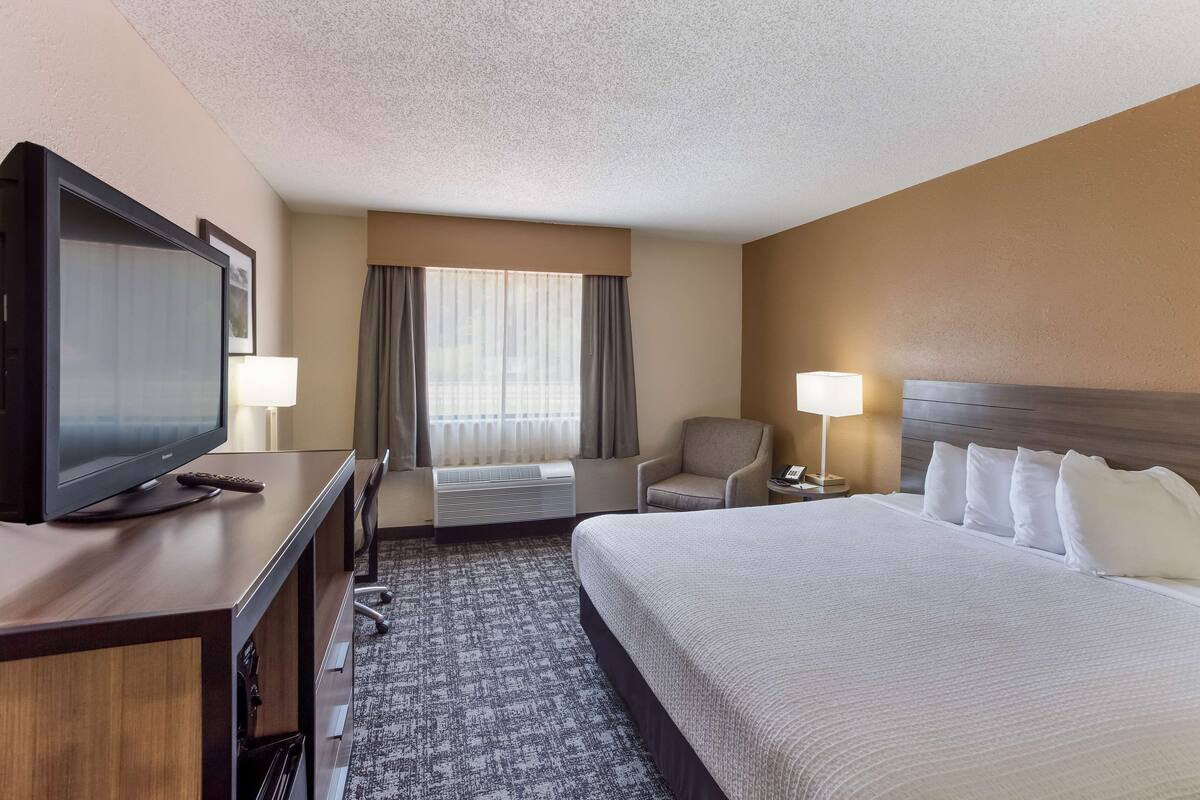 A hotel room with a large bed, a TV on a wooden desk, a chair, a desk lamp, and a window with curtains.