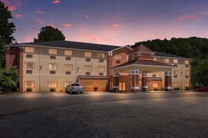 Un hotel de varios pisos con una entrada central, estacionamiento y un hermoso paisaje de árboles y un cielo al atardecer.