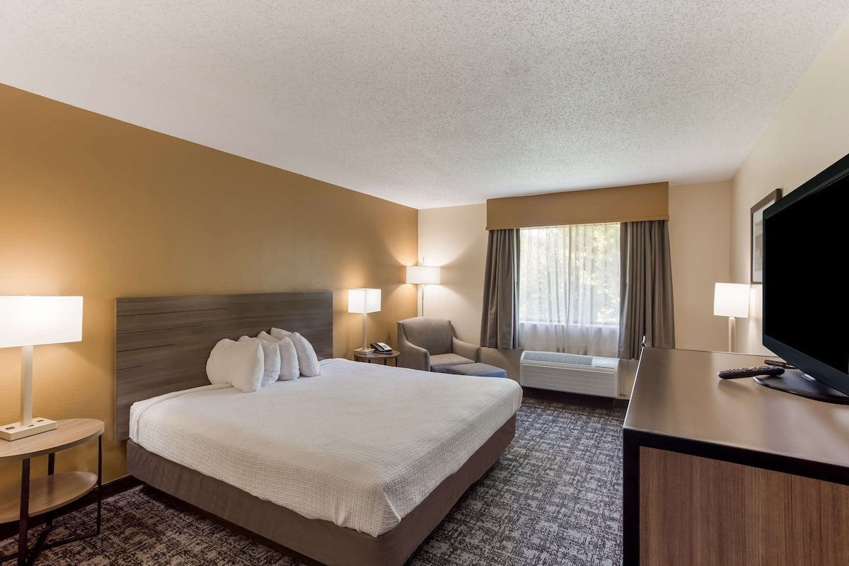 A hotel room with a large bed, two bedside tables with lamps, a chair, a desk with a TV, and a window with curtains.
