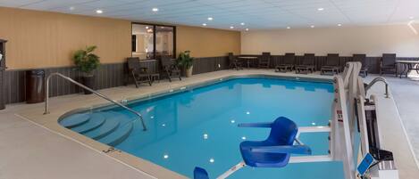 An indoor swimming pool with a clear blue water, surrounded by lounge chairs and a seating area with tables.