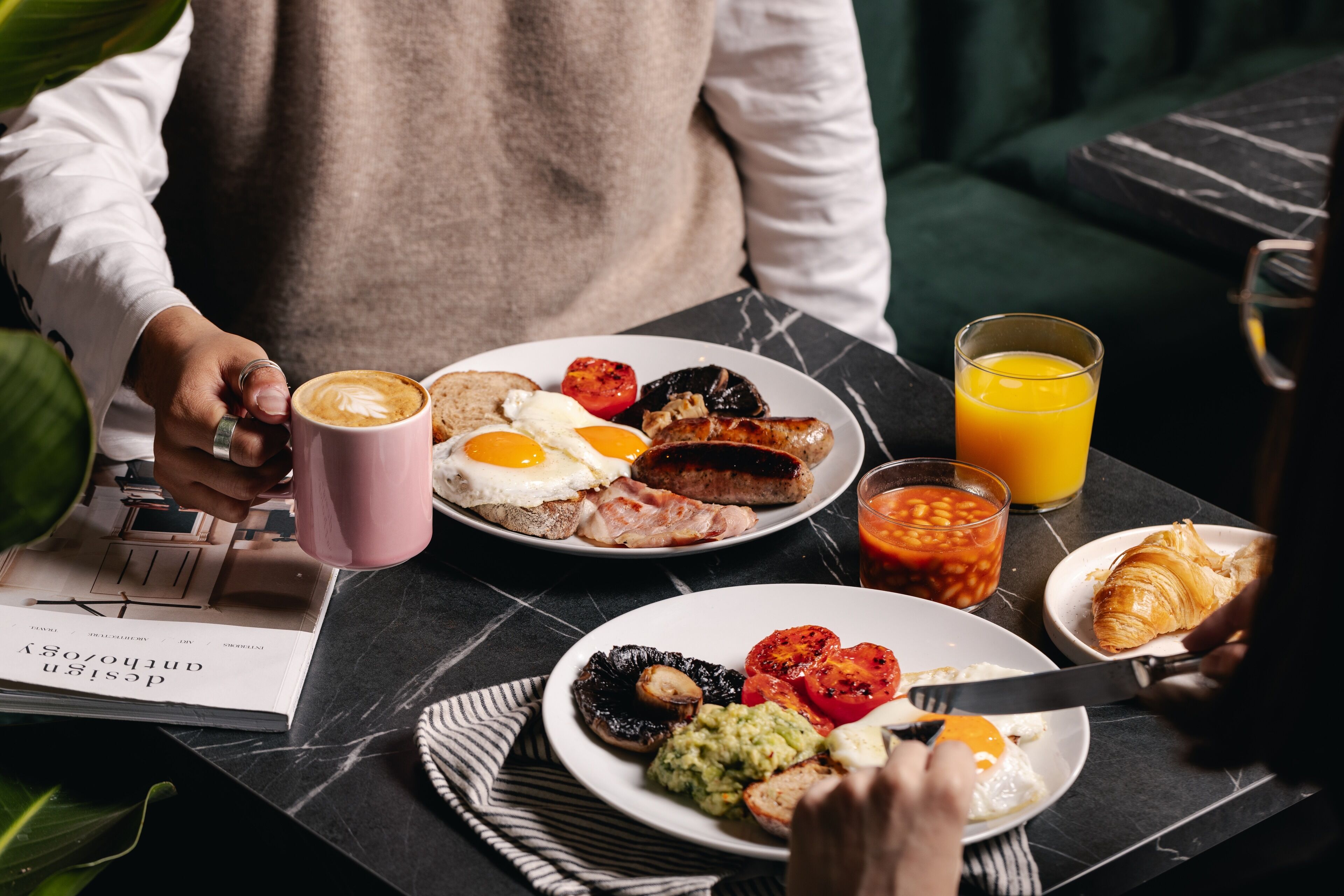 Mesa de café da manhã com diversos alimentos, incluindo ovos, salsichas, torradas e um croissant, além de um copo de suco de laranja e uma xícara de café.