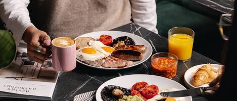 Mesa de café da manhã com diversos alimentos, incluindo ovos, salsichas, torradas e um croissant, além de um copo de suco de laranja e uma xícara de café.